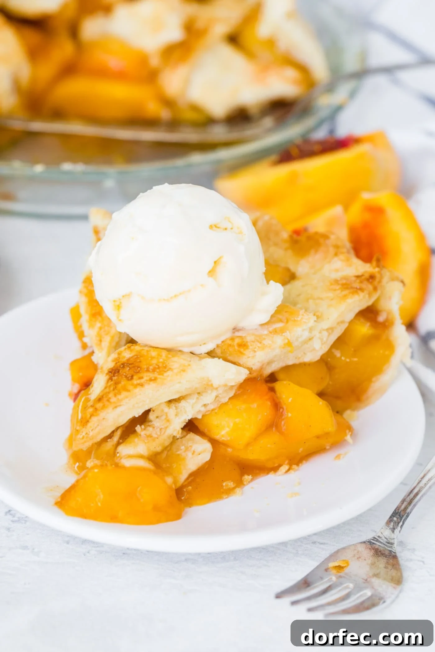 A photo of a slice of fresh peach pie with lattice crust on a white plate with a scoop of vanilla ice cream on top.