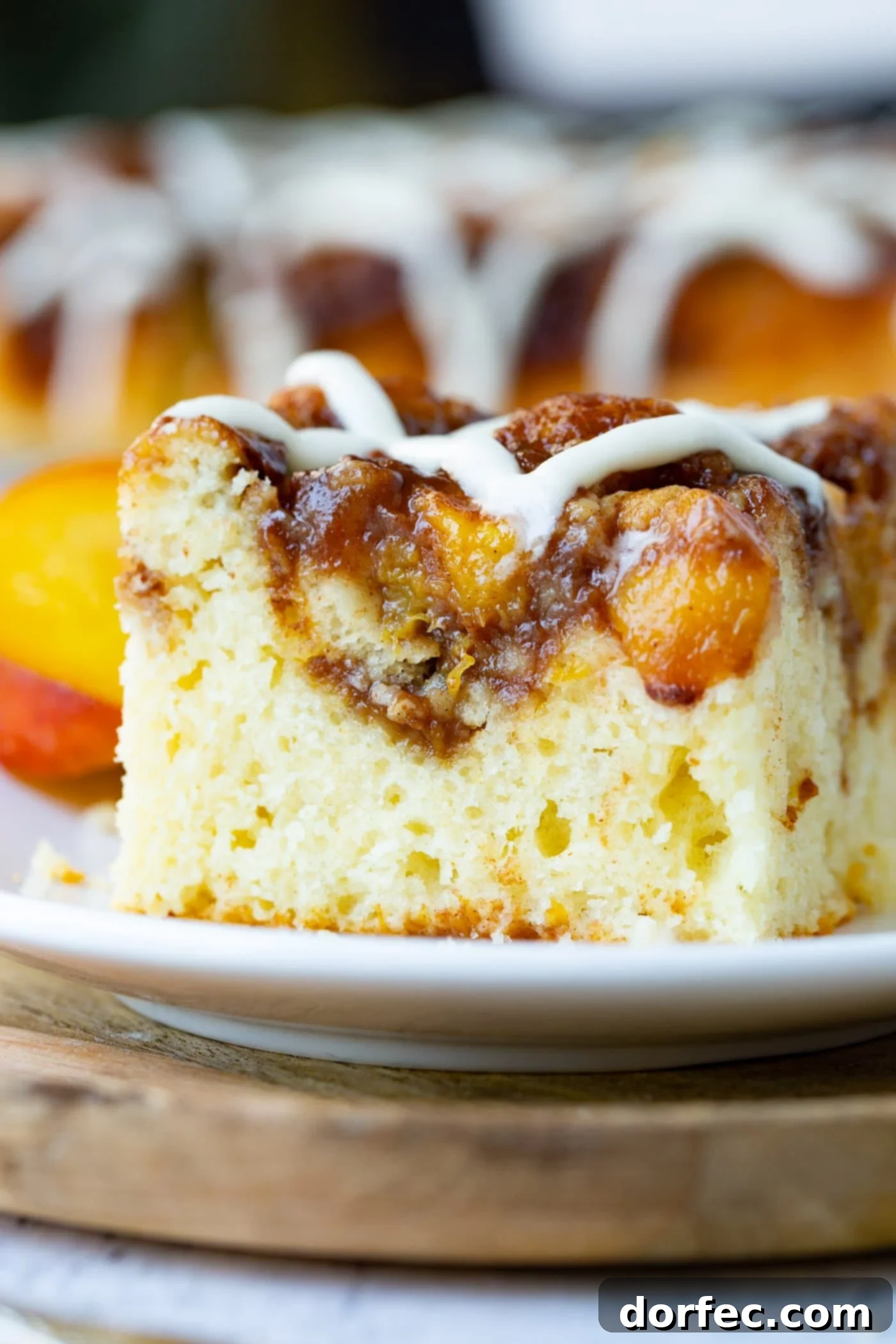 A slice of easy peach sour cream coffee cake, topped with a rich brown sugar peach topping and a sweet glaze, resting on a small white plate atop a rustic wooden board.