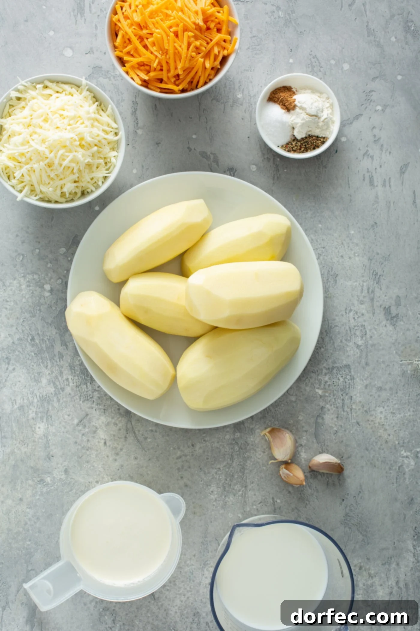 A bowl of six peeled potatoes. There are small containers of grated cheese, 