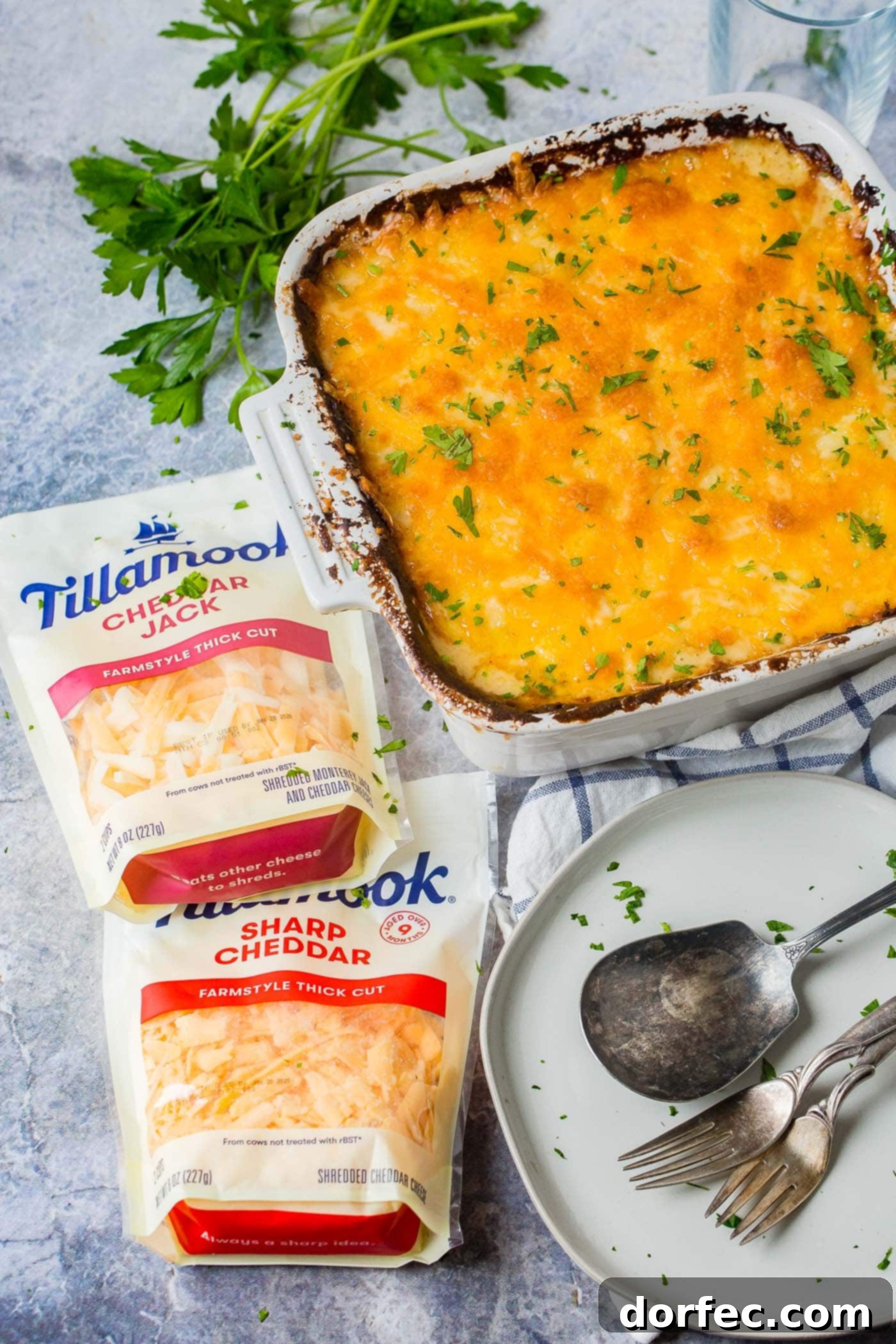 The Ultimate Cheesy Potato Casserole 4 A baking dish filled with golden-brown cheesy potato casserole, flanked by two bags of Tillamook shredded cheese and a plate with cutlery, emphasizing the quality ingredients.