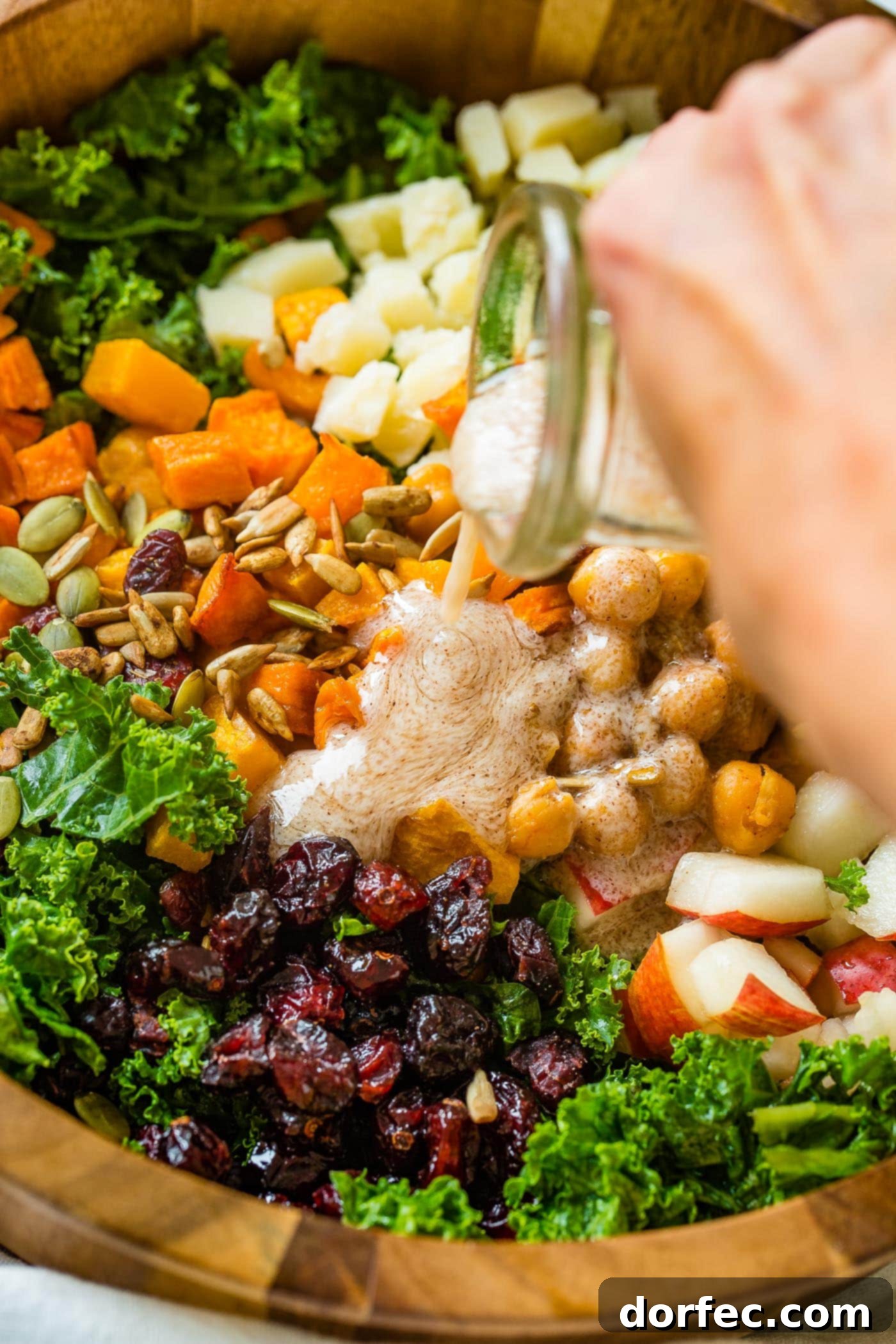 Golden Roasted Butternut & Kale Power Salad 5 A photo of dressing being poured on a large wooden bowl full of kale salad topped with roasted butternut squash, craisins, chickpeas, pepitas, and gouda cheese.