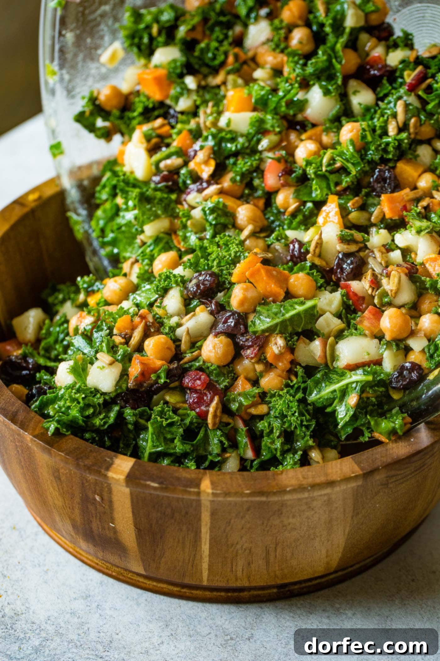 Golden Roasted Butternut & Kale Power Salad 6 A photo of a large wooden bowl full of kale salad topped with roasted butternut squash, craisins, chickpeas, pepitas, and gouda cheese all being tossed.