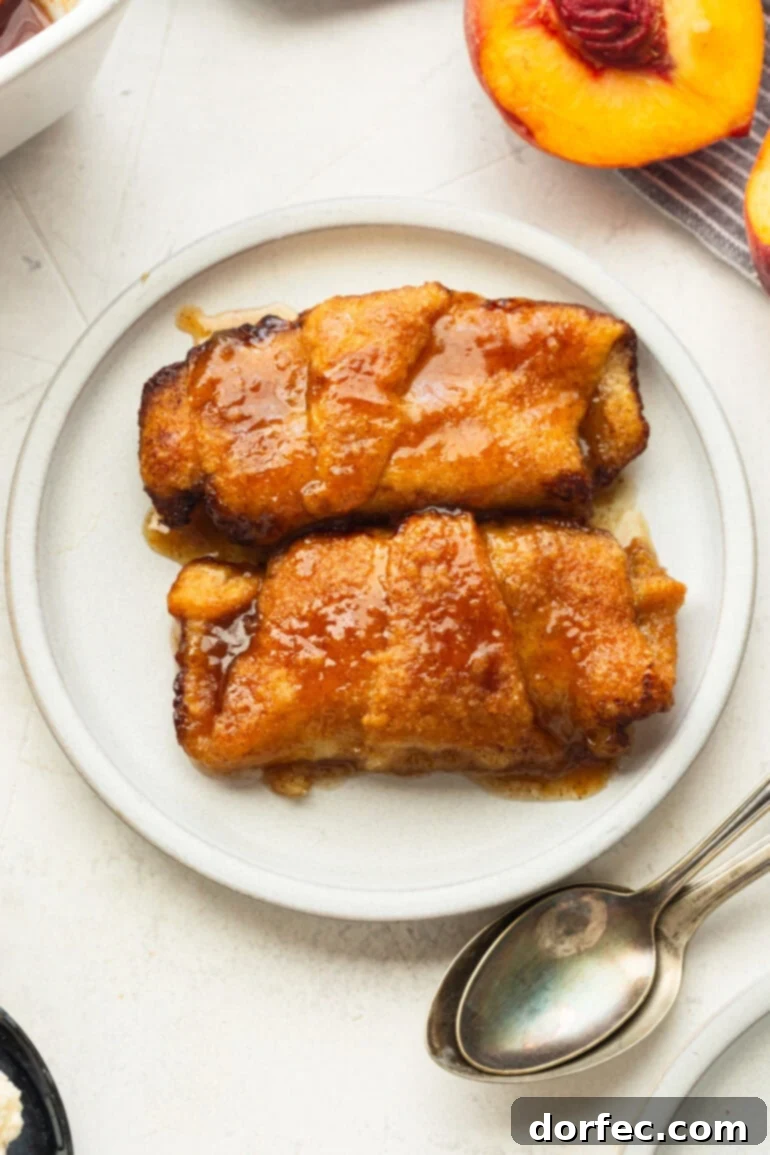 Two golden baked peach dumplings with crescent rolls, glistening with sauce, sitting invitingly on a small white dessert plate, ready to be enjoyed.
