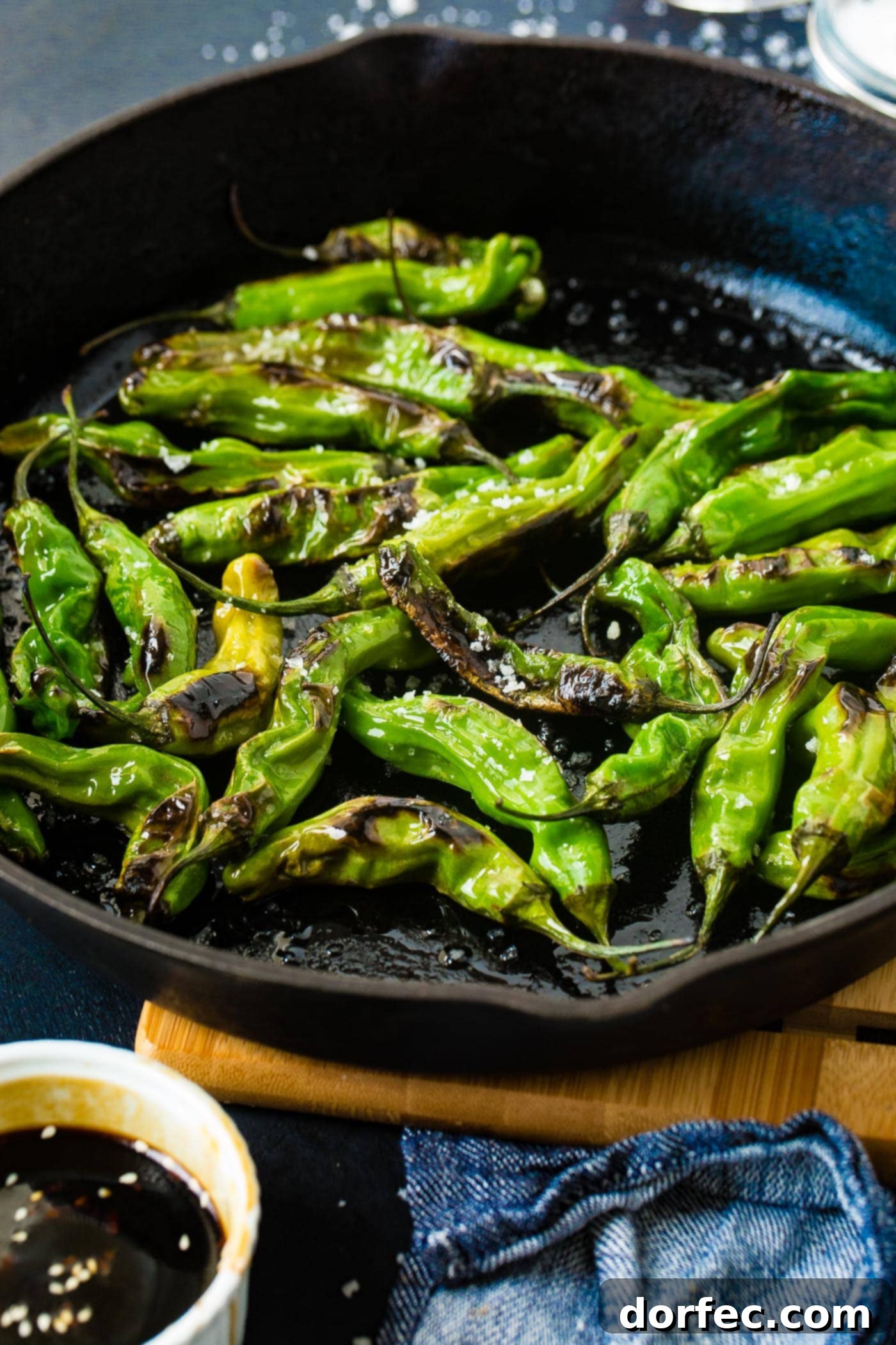 a black cast iron pan with bright green blistered shishito peppers and flaked sea salt