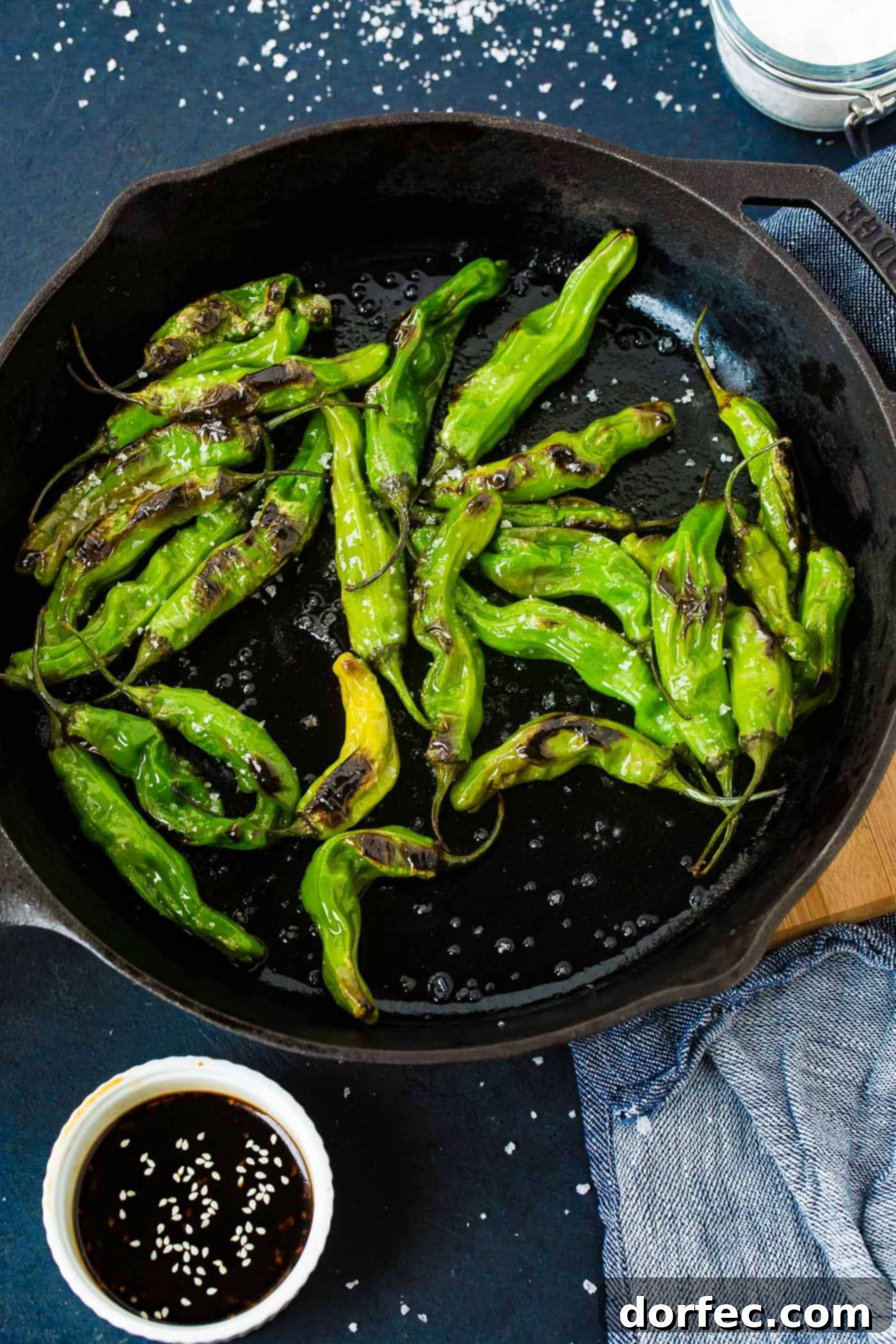 a black cast iron pan with bright green blistered shishito peppers and flaked sea salt