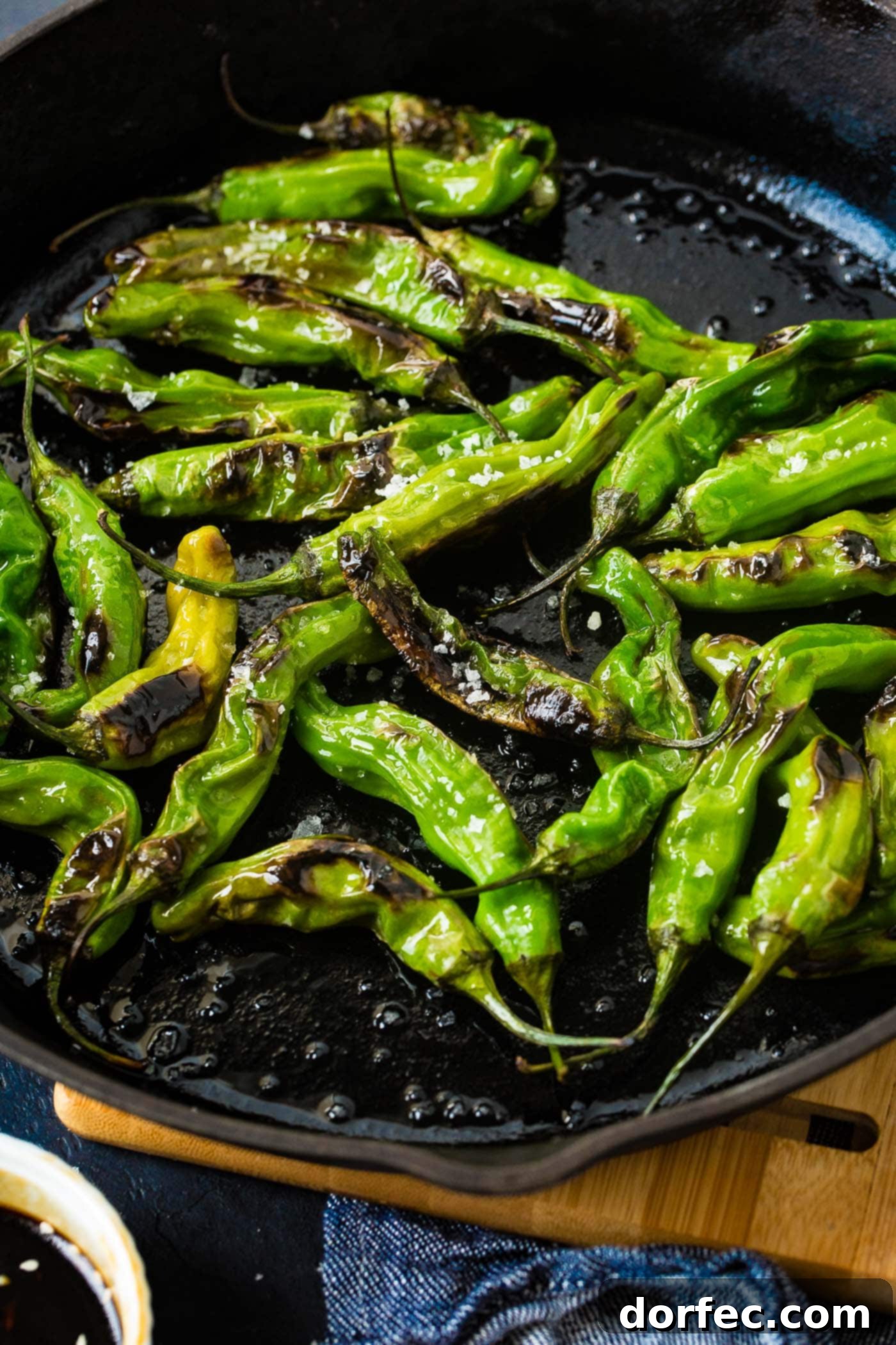 a black cast iron pan with bright green blistered shishito peppers and flaked sea salt
