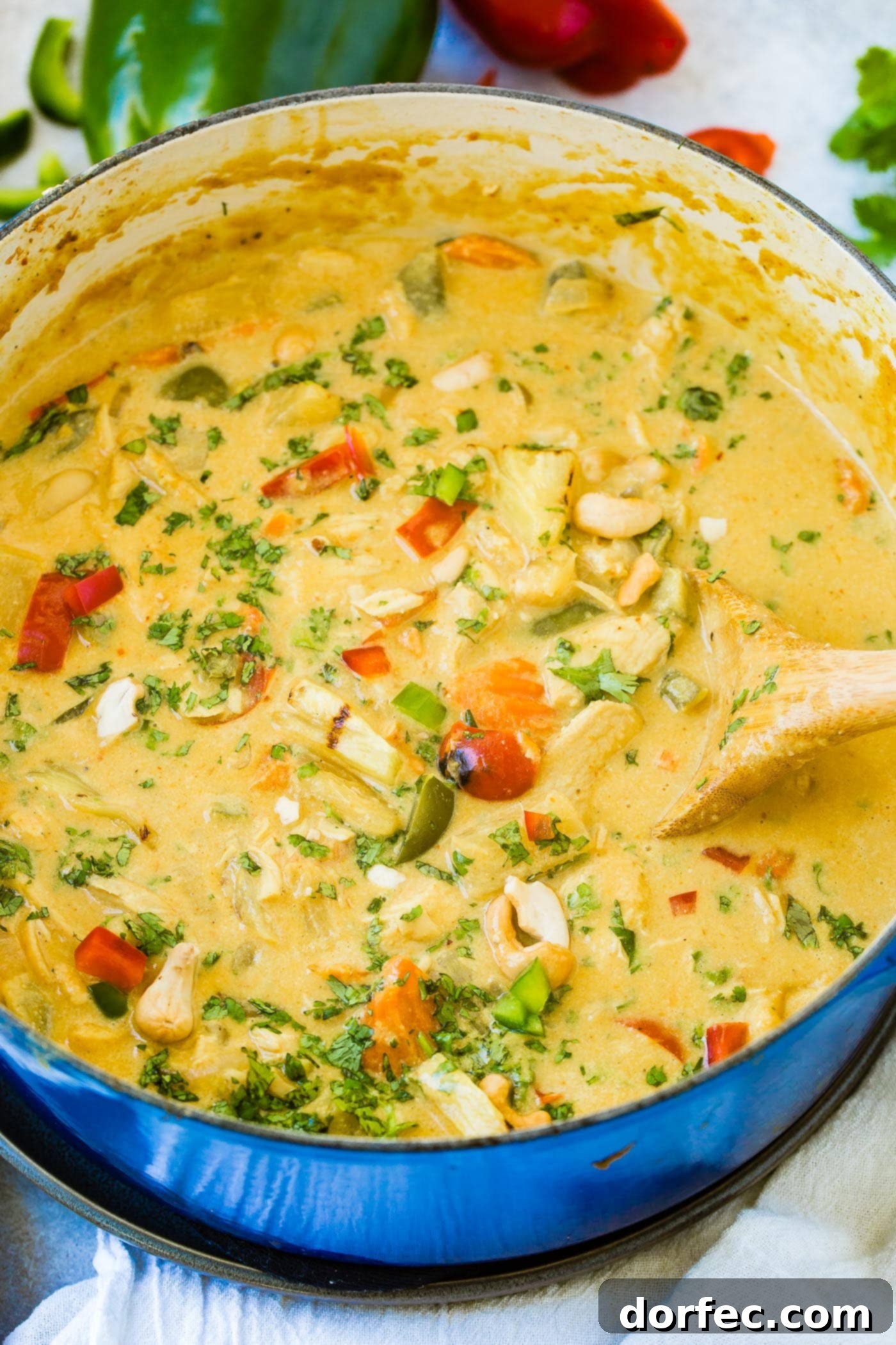 A close-up view of a large Dutch oven filled with simmering creamy pineapple coconut chicken curry, rich with colorful bell peppers, tender onions, and chicken pieces. A scattering of crunchy cashews rests on top, inviting a spoonful.