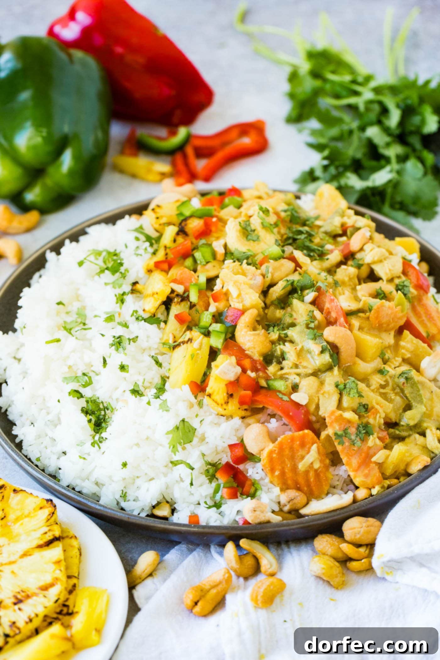 A vibrant and inviting plate of creamy pineapple coconut chicken curry, expertly plated over a bed of fluffy jasmine white rice. The curry is richly textured with colorful bell peppers, tender onions, and a generous sprinkle of salty cashews, while a perfectly grilled pineapple slice adds a touch of gourmet flair and tropical sweetness.