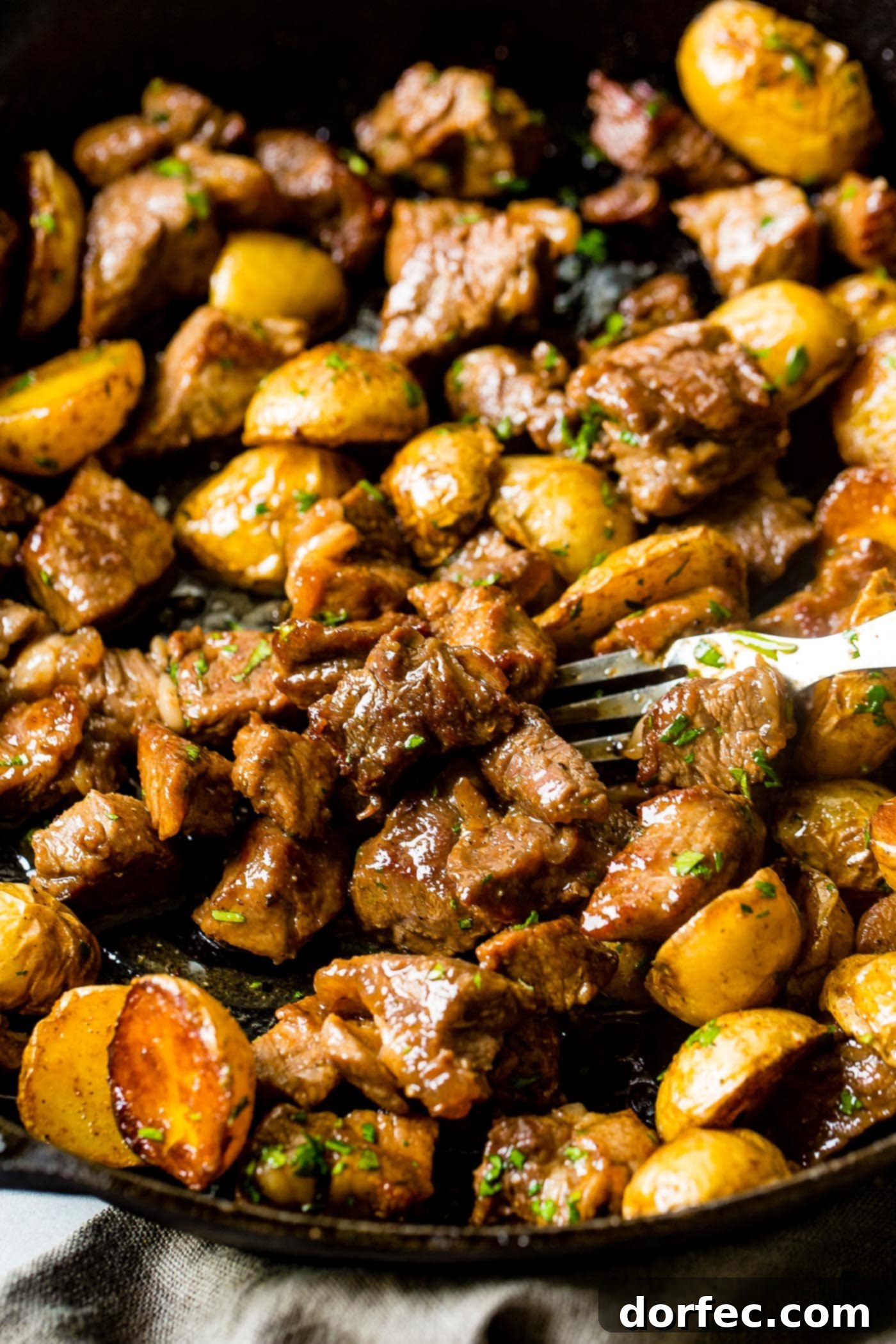 A photo of a cast iron skillet full of golden halved potatoes and beautifully seared steak, garnished with fresh herbs.