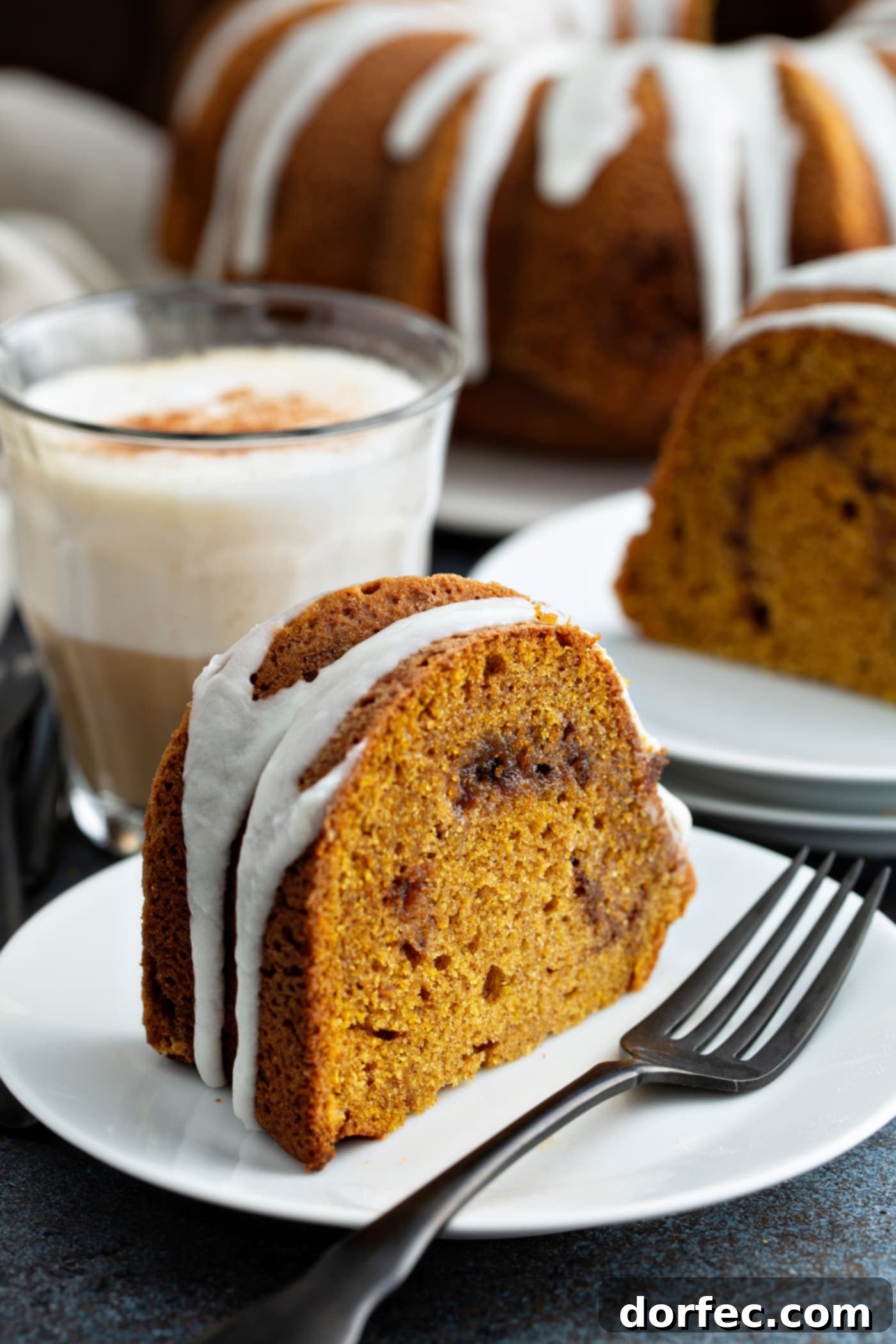 Sour Cream Cinnamon Swirl Pumpkin Bundt Cake A slice of sour cream cinnamon swirl pumpkin bundt cake on a dessert plate with a fork on the plate next to the cake. The cake has a drizzle of white glaze on top. another slice of cake on a dessert plate, and the rest of the bundt cake on a cake platter are in the background.