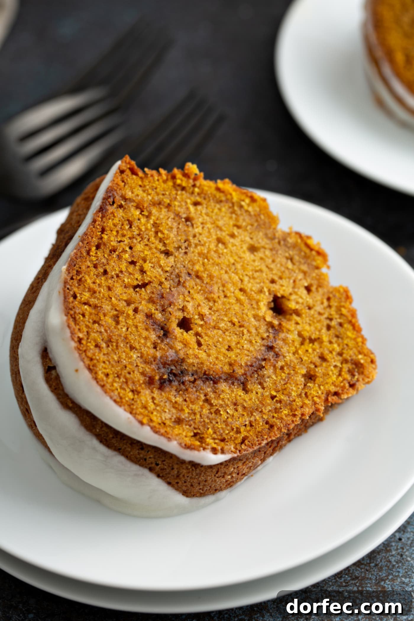 Sour Cream Cinnamon Swirl Pumpkin Bundt Cake A slice of sour cream cinnamon swirl pumpkin cake on a dessert plate. The cake is drizzled with glaze and the cinnamon swirl in the middle is visible. A fork is next to the plate and another dessert plate is in the background.