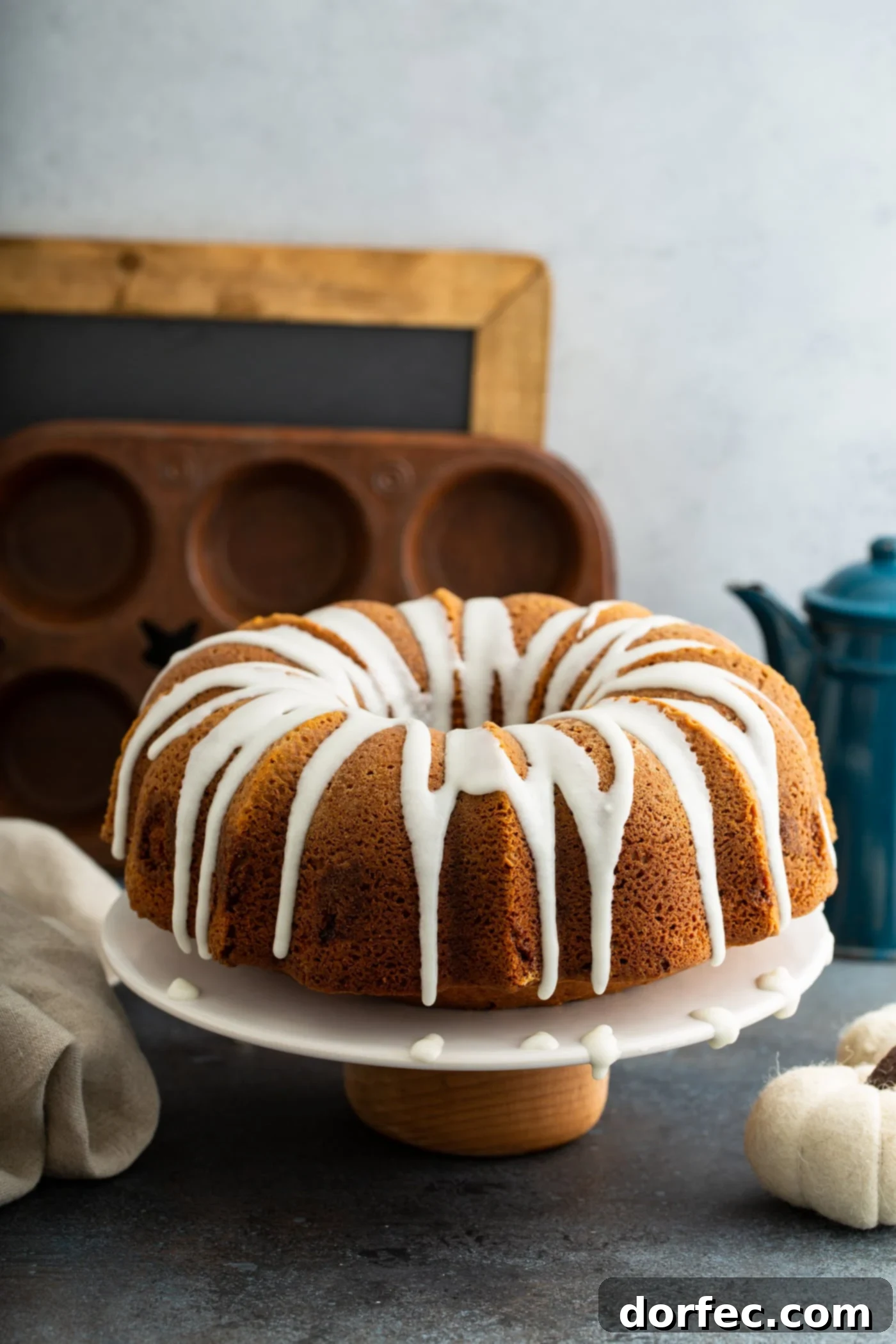 Sour Cream Cinnamon Swirl Pumpkin Bundt Cake A sour cream cinnamon swirled pumpkin bundt cake with white glaze drizzled over the top. The cake is on a white cake plate and a muffin tin, chalk board, and coffee pot are in the background.