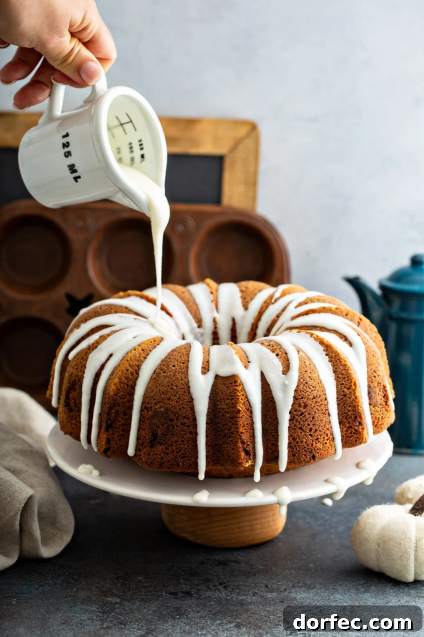Sour Cream Cinnamon Swirl Pumpkin Bundt Cake White glaze being drizzled from a glass measuring cup over the top of a sour cream cinnamon swirled pumpkin bundt cake. A muffin tin, a chalk board, and a blue coffee pot are in the background.