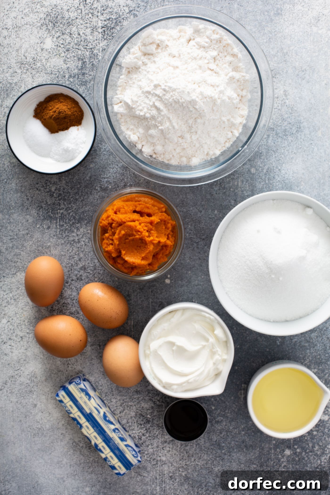 Sour Cream Cinnamon Swirl Pumpkin Bundt Cake Small containers with ingredients for sour cream cinnamon swirl pumpkin bundt cake. There are containers of flour, sugar, sour cream, butter, pumpkin, and cinnamon. there are also four brown eggs on the table.
