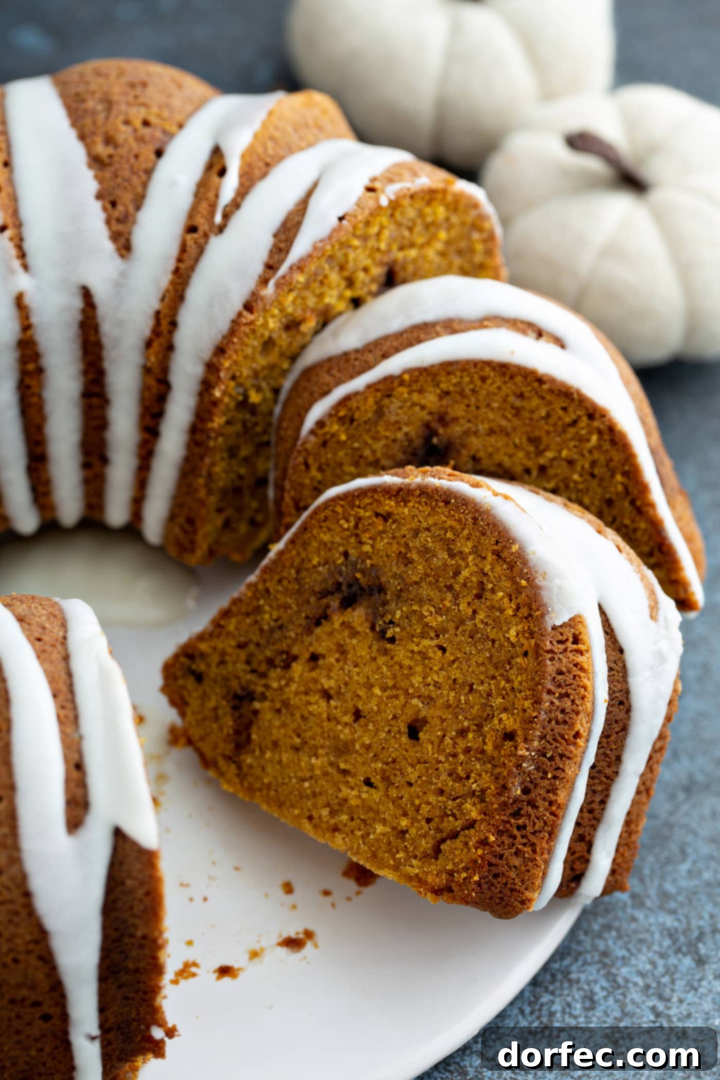 Sour Cream Cinnamon Swirl Pumpkin Bundt Cake A sour cream cinnamon swirl pumpkin bundt cake on a cake platter. A couple of pieces of cake have been sliced. The cake has a white drizzled frosting on top.