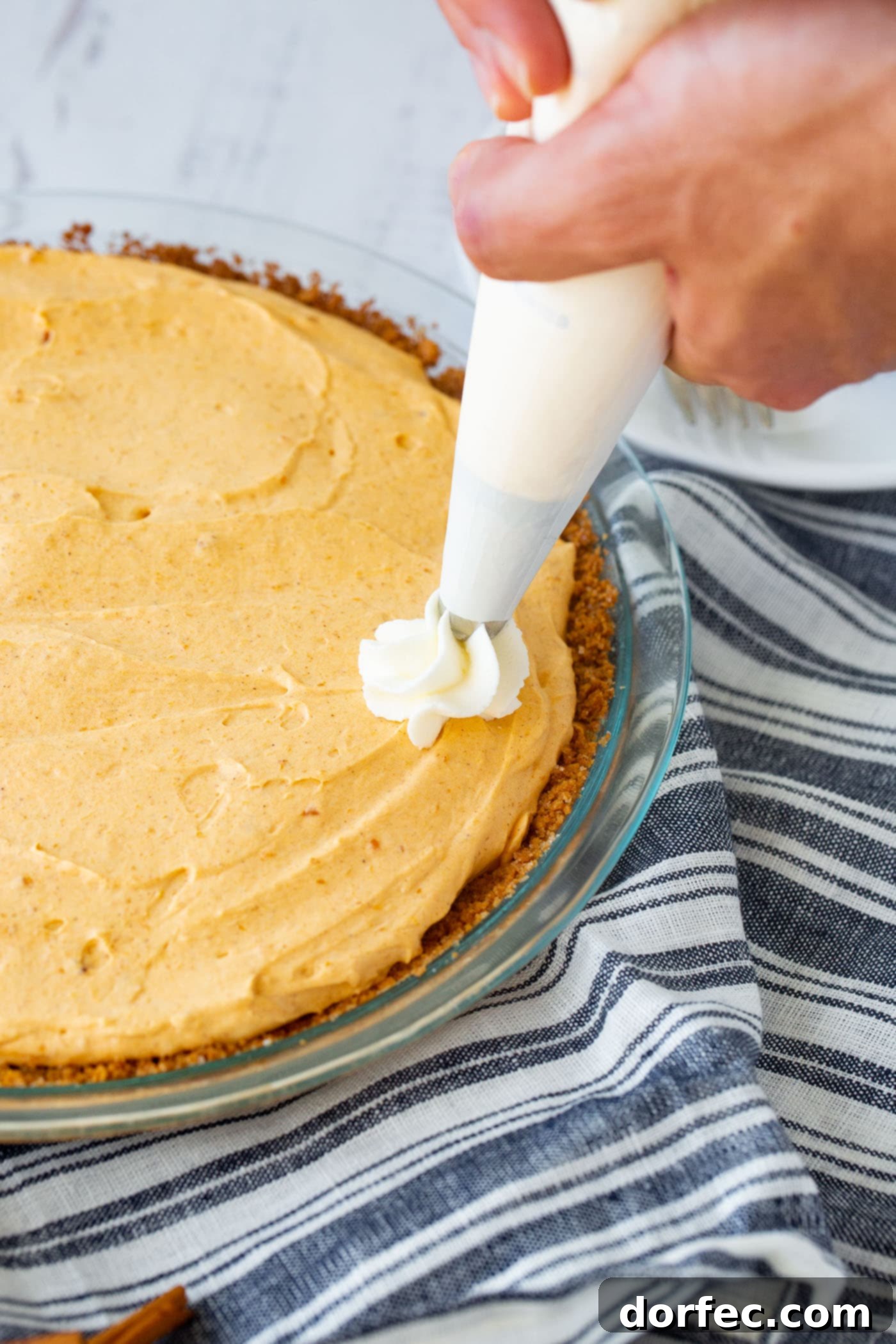 Marshmallow No Bake Pumpkin Pie A photo of a no bake pumpkin pie with beautiful swirls of whipped cream being meticulously piped on top, adding the finishing touch before serving.