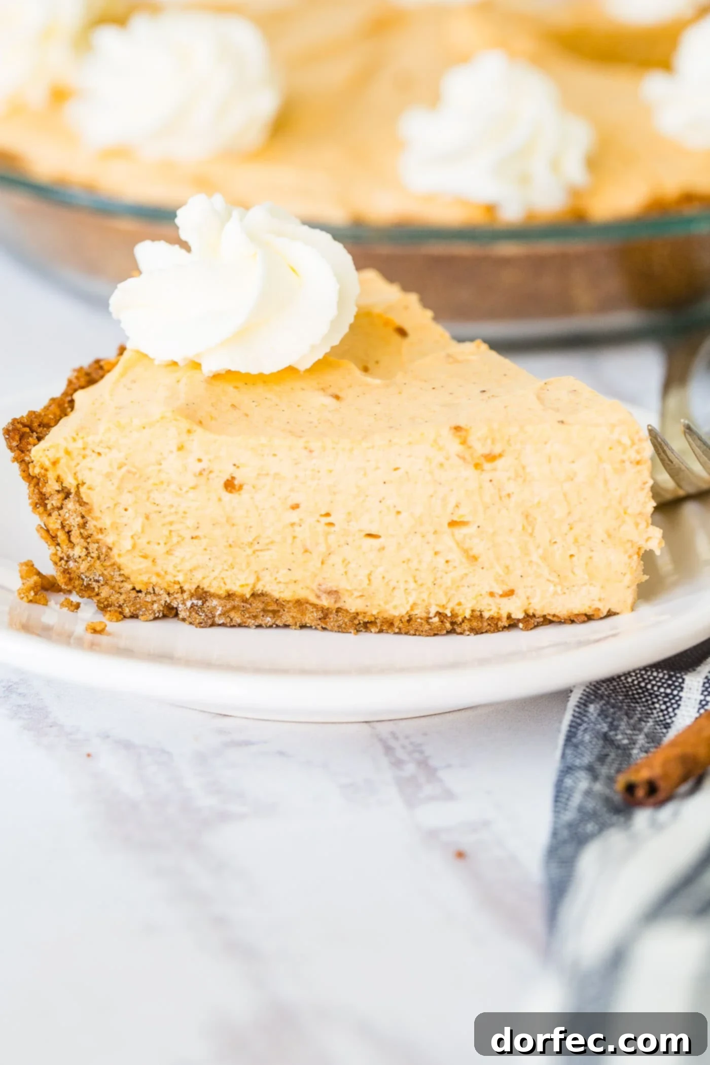 Marshmallow No Bake Pumpkin Pie A photo of a slice of no bake pumpkin pie on a white plate with a swirl of whipped cream on top, expertly plated and ready to be served.