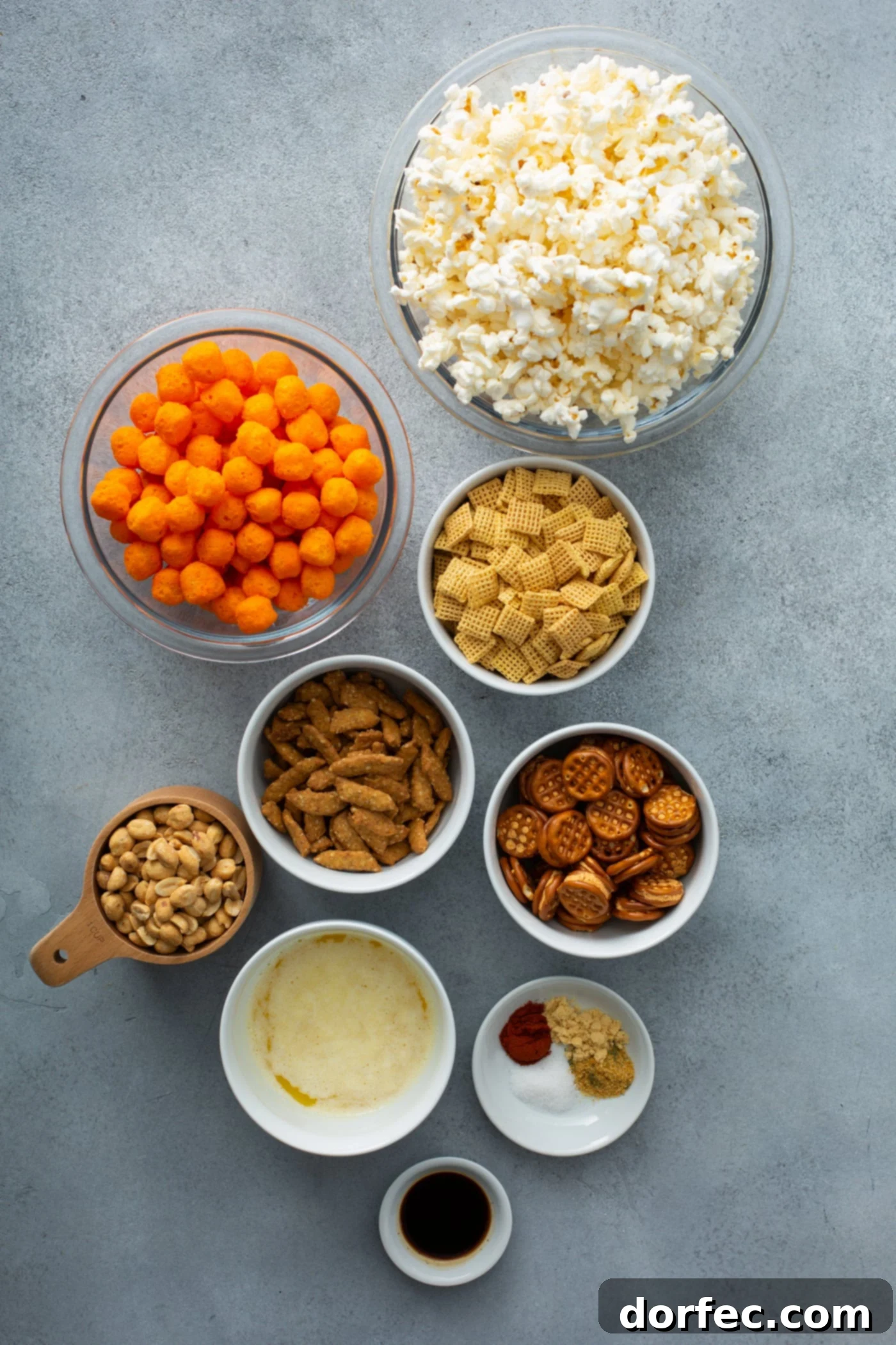 Various containers filled with the components for a savory snack mix. Ingredients like popcorn, cheese balls, pretzels, sesame sticks, peanuts, and a selection of spices are displayed, ready for assembly.