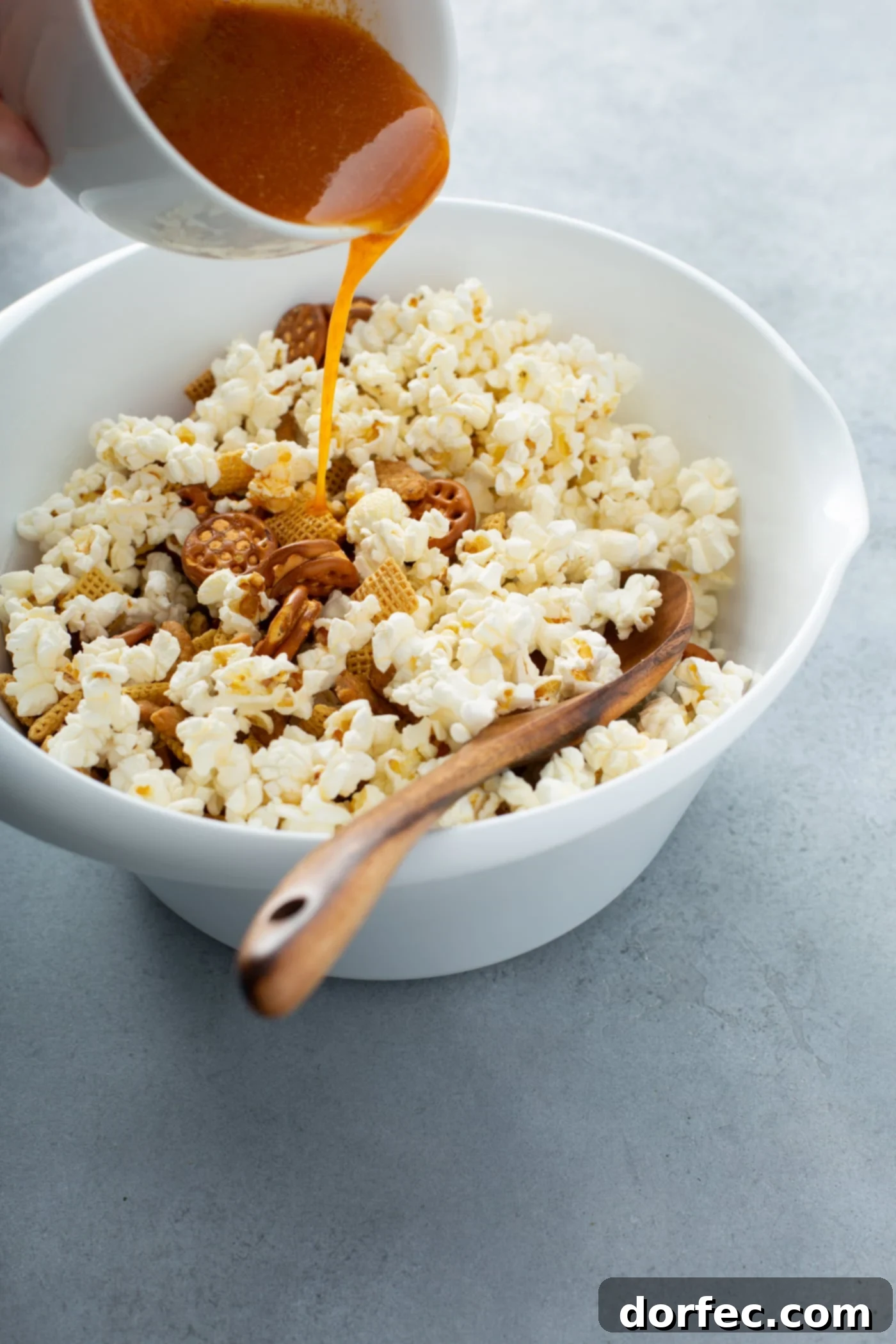 A large bowl containing popcorn and other dry snack mix ingredients. Melted butter and a blend of spices are being carefully poured over the mix, ready for tossing and coating.