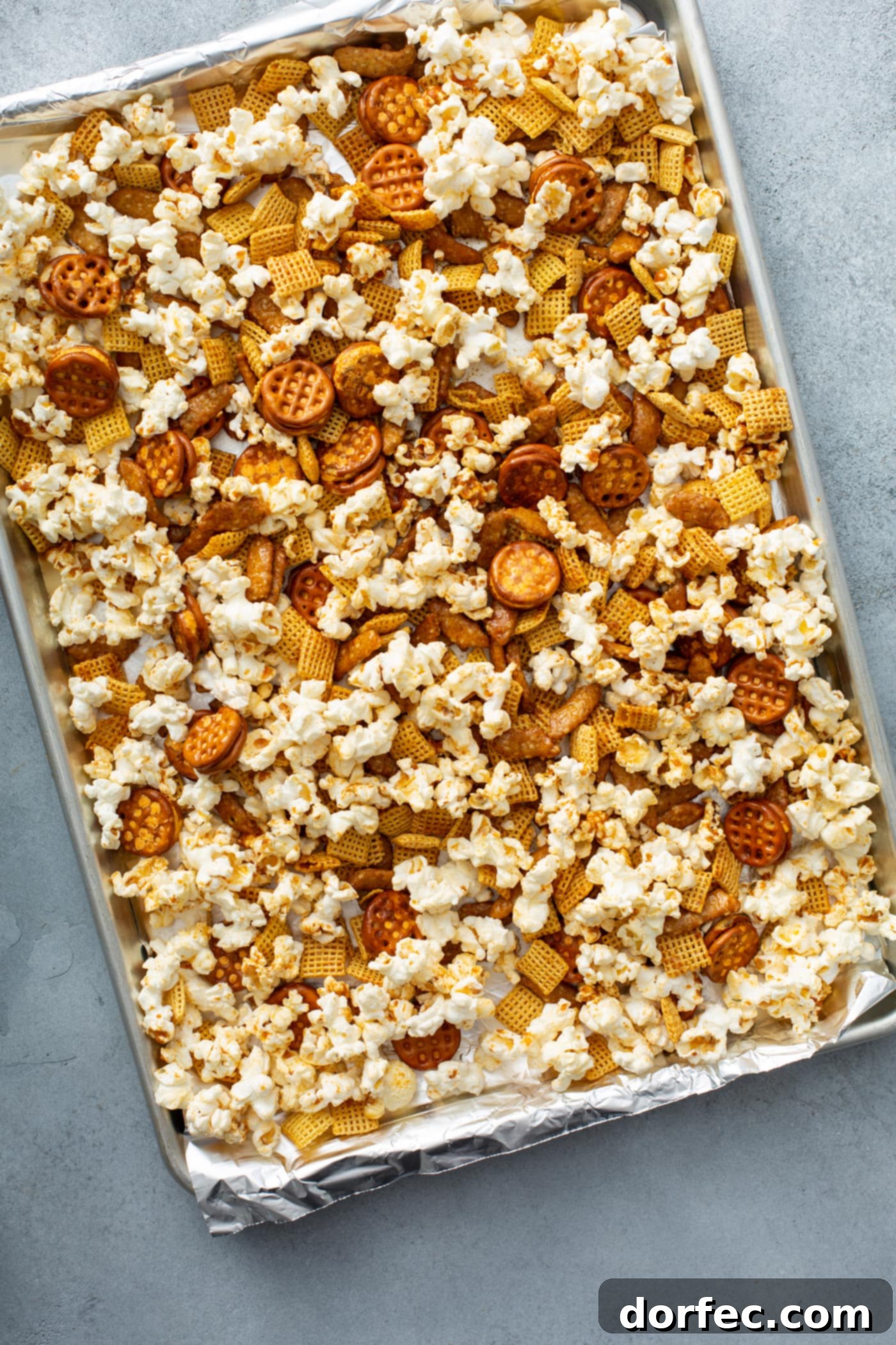 A baking sheet generously filled with a single layer of popcorn, Chex cereal, pretzels, and sesame sticks, freshly baked and ready to cool.