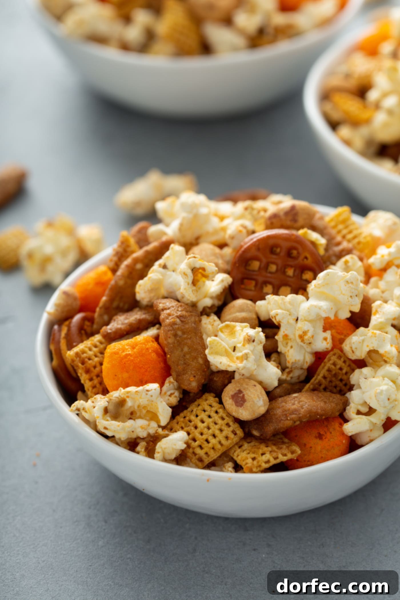 A large bowl of savory March Madness snack mix, featuring popcorn, peanuts, Chex, pretzels, and sesame sticks, with additional bowls blurred in the background, set for a game day celebration.