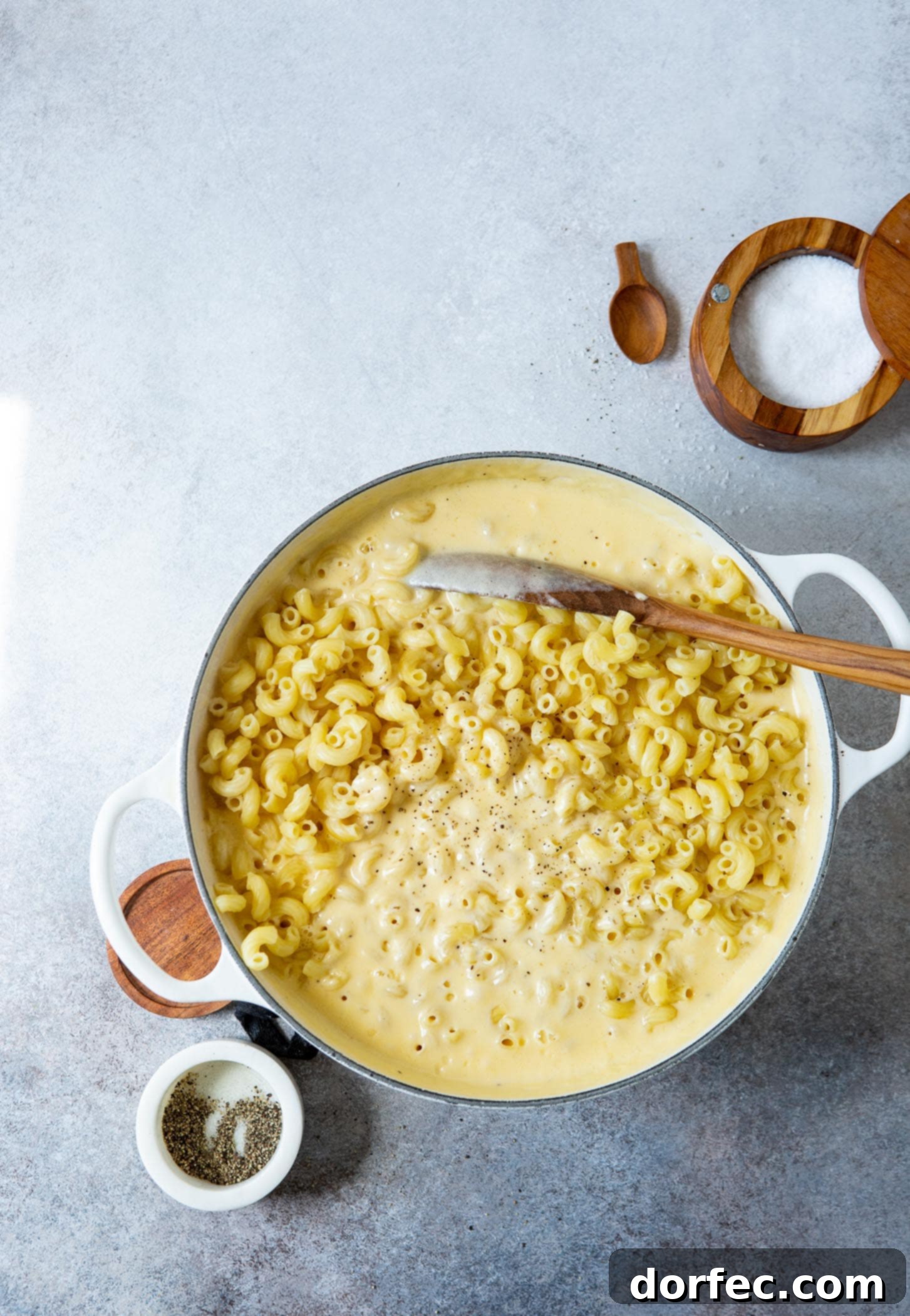 Best Macaroni and Cheese Ever A pan with macaroni, cheese sauce and a wooden spoon. A container of salt is in the background.