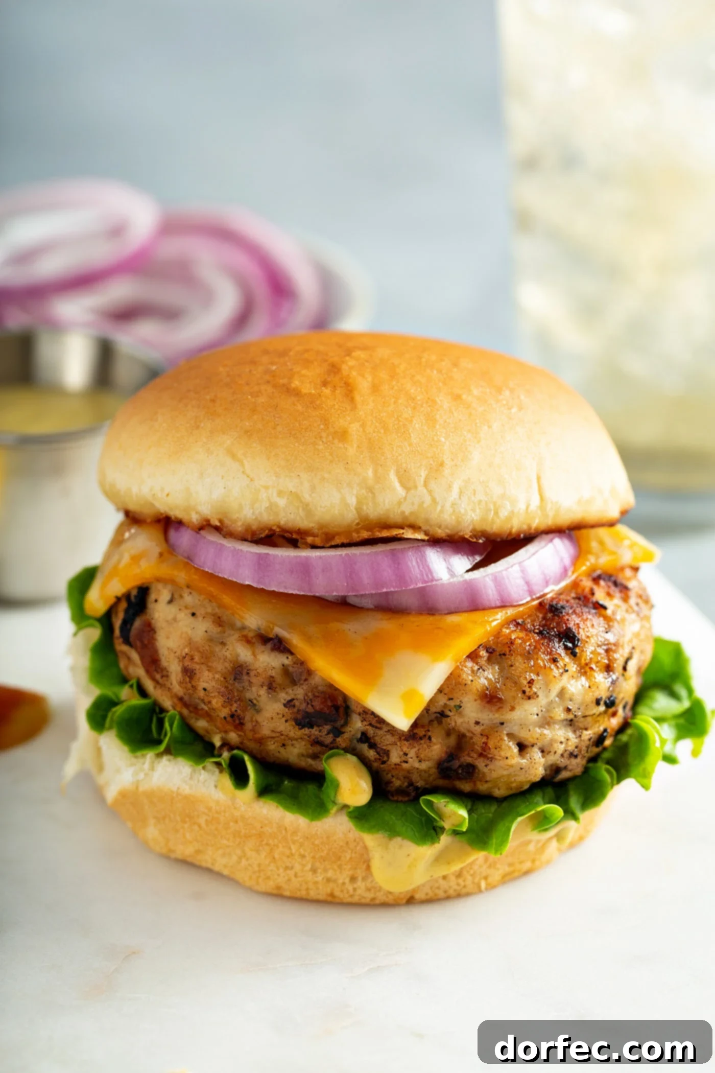 A maple mustard chicken burger on a bun with lettuce, cheese and onions. There is a plate of sliced red onions in the background.