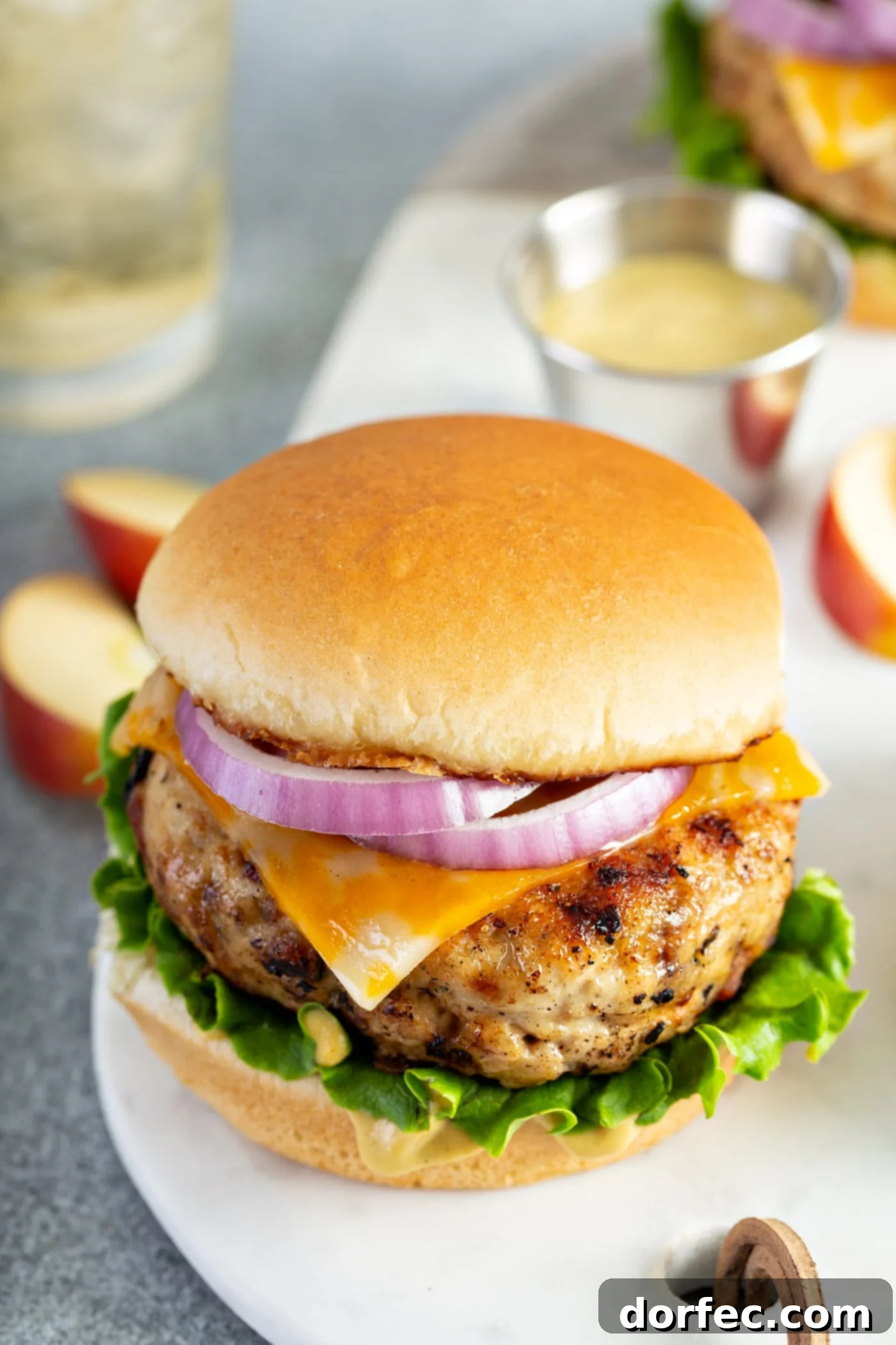 A chicken burger on a bun with lettuce, cheese and red onion. There are apple slices and a glass of apple juice in the background.