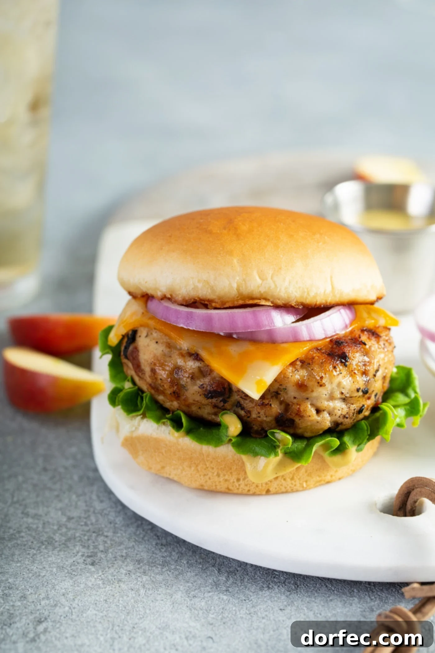 A chicken burger on a white plate. The burger is on a bun wing lettuce, cheese, and red onion. There are two slices of apple beside the plate.