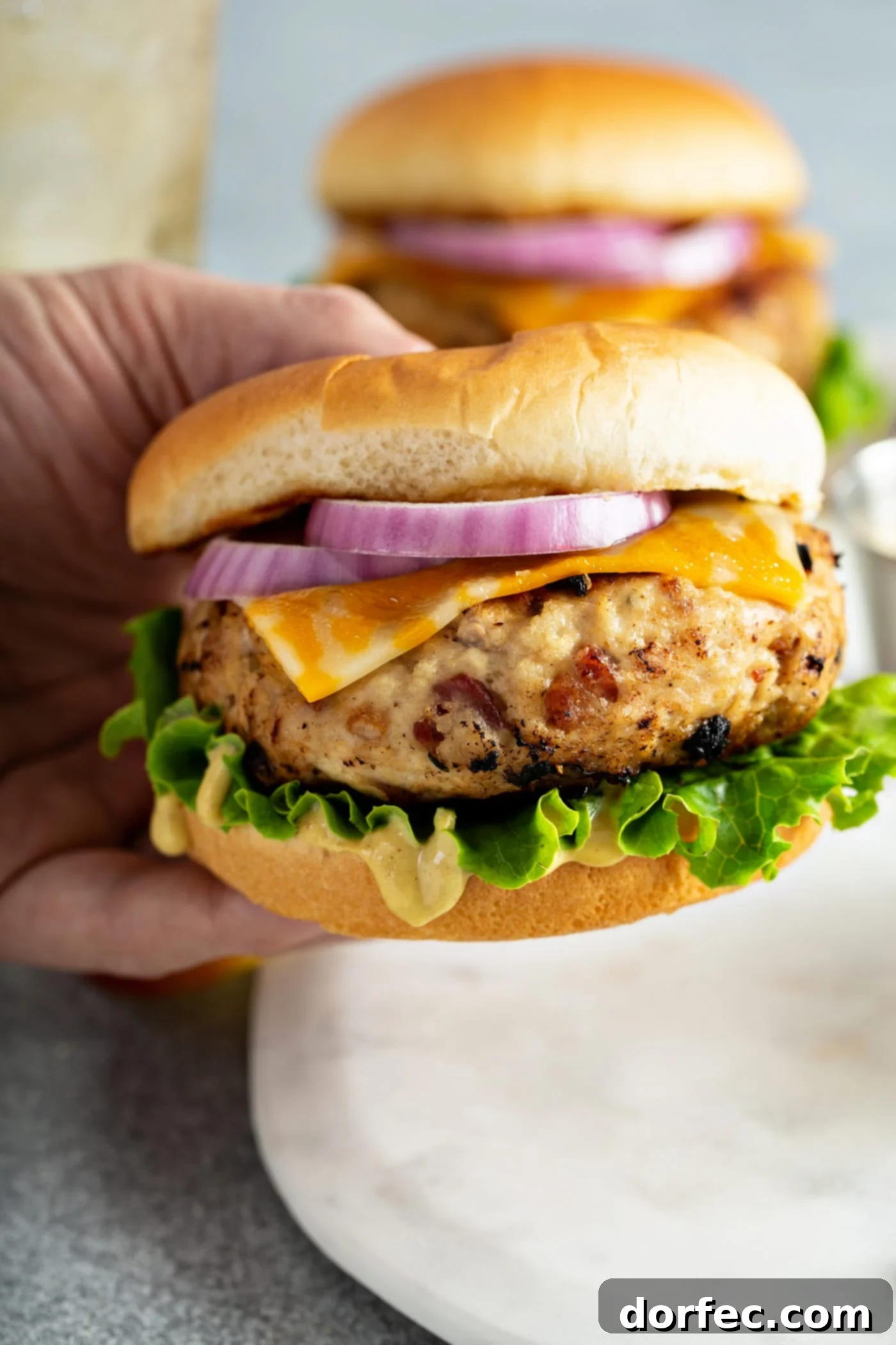 A chicken burger being held in a hand. The burger is in a bun with lettuce, cheese, and sliced red onion.