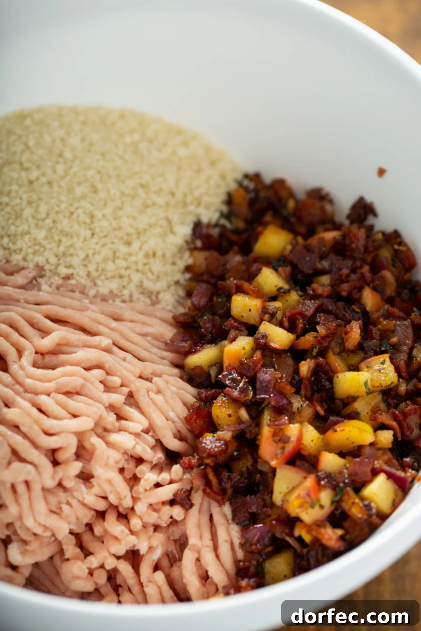 A plate with a scoop of bread crumbs in one area, ground chicken next to the bread crumbs and the cooked bacon, apple and spice mixture next to them.