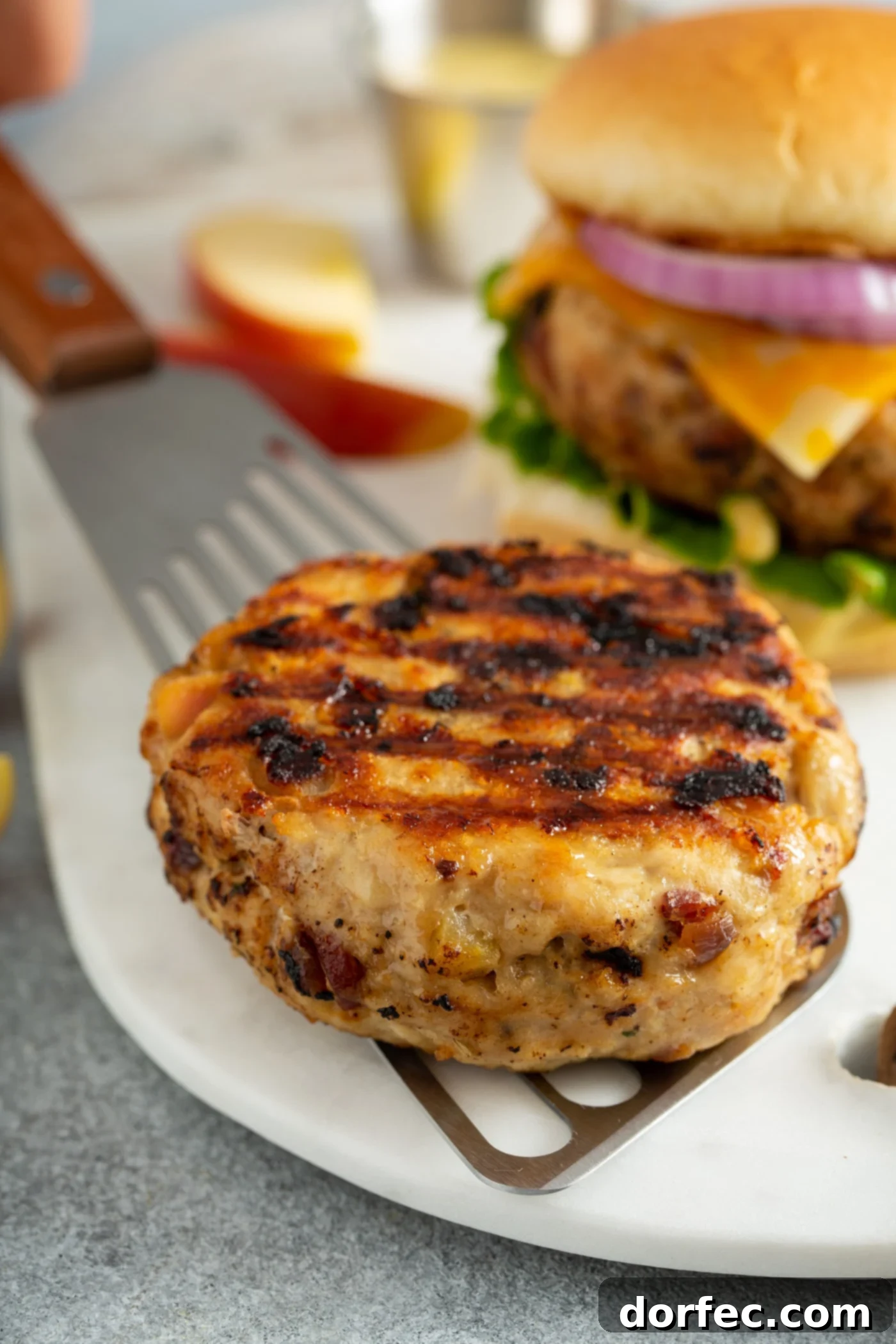 A grilled chicken burger patty being placed on a white plate with a spatula. There are apple slices and another chicken burger in the background.. 