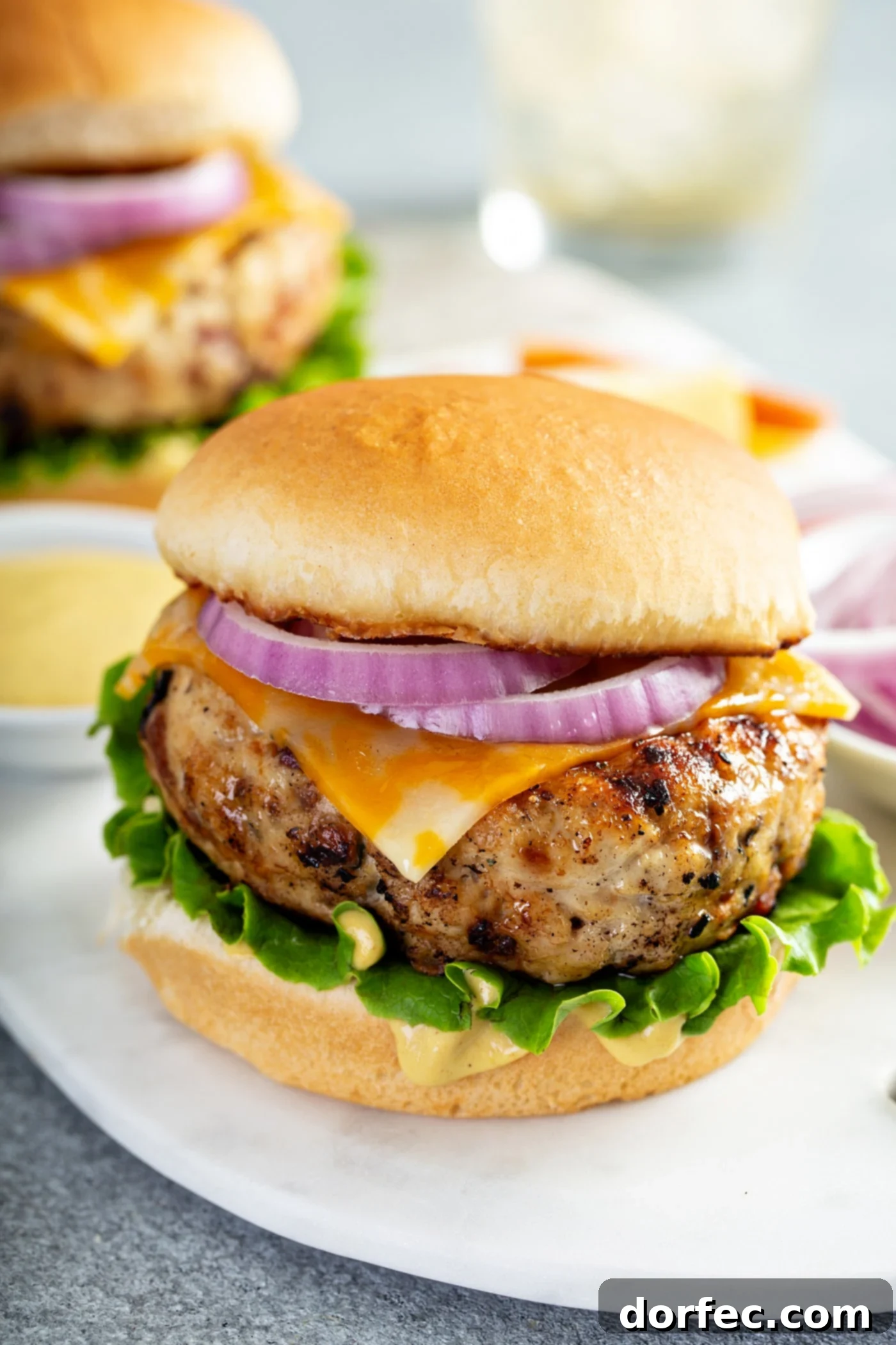 A chicken burger on a bun with lettuce, cheese, and red onion. There is another chicken burger in the background and small containers of bread crumbs, and red onions.