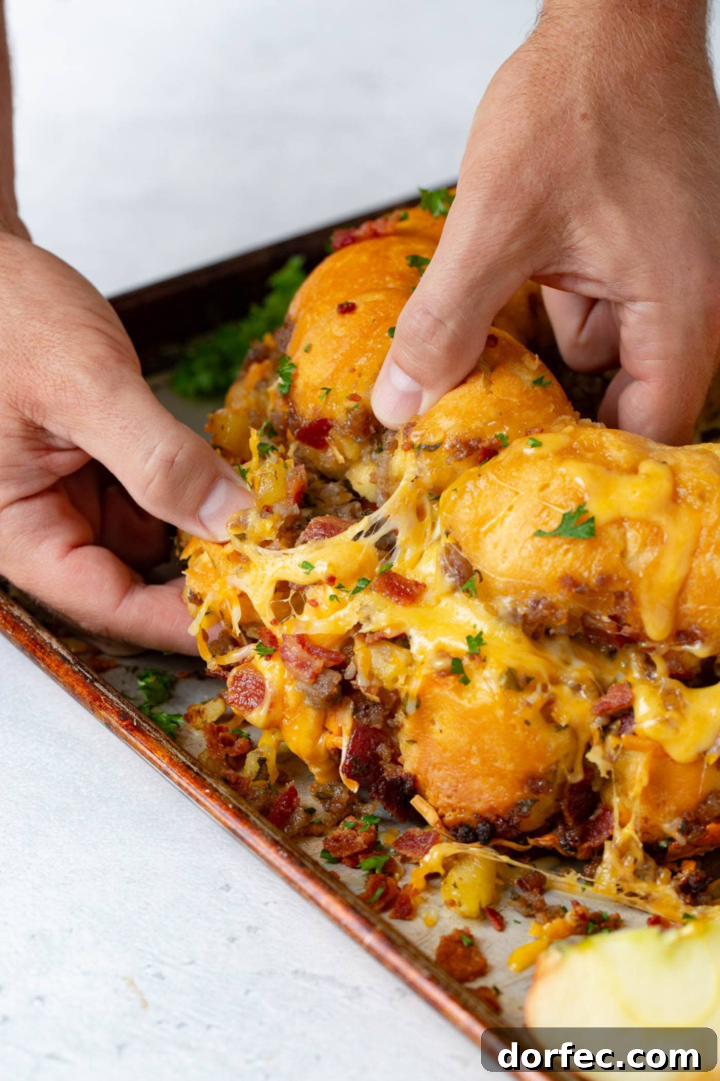 Breakfast Sausage and Apple Cheesy Pull Apart A hand pulling apart a section of Breakfast Cheesy Pull Apart Bread, showcasing the melted cheese, savory breakfast sausage, crispy bacon, and soft apple chunks within.