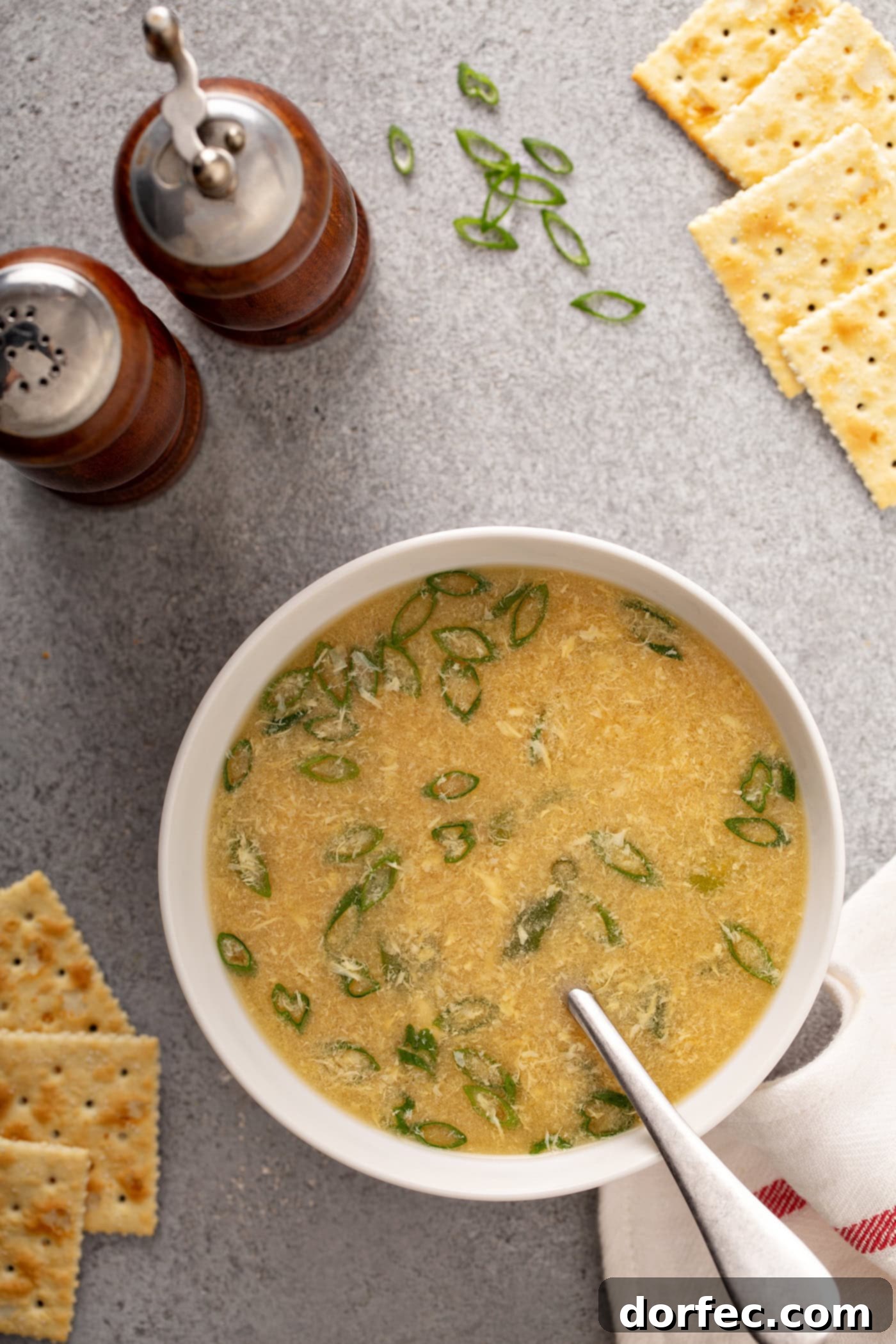 Easy Egg Drop Soup A table top with wooden salt and pepper shakers, saltine crackers, sliced green onions and a bowl of egg drop soup with a spoon in it. You can see the bits of egg and sliced green onions in the soup.