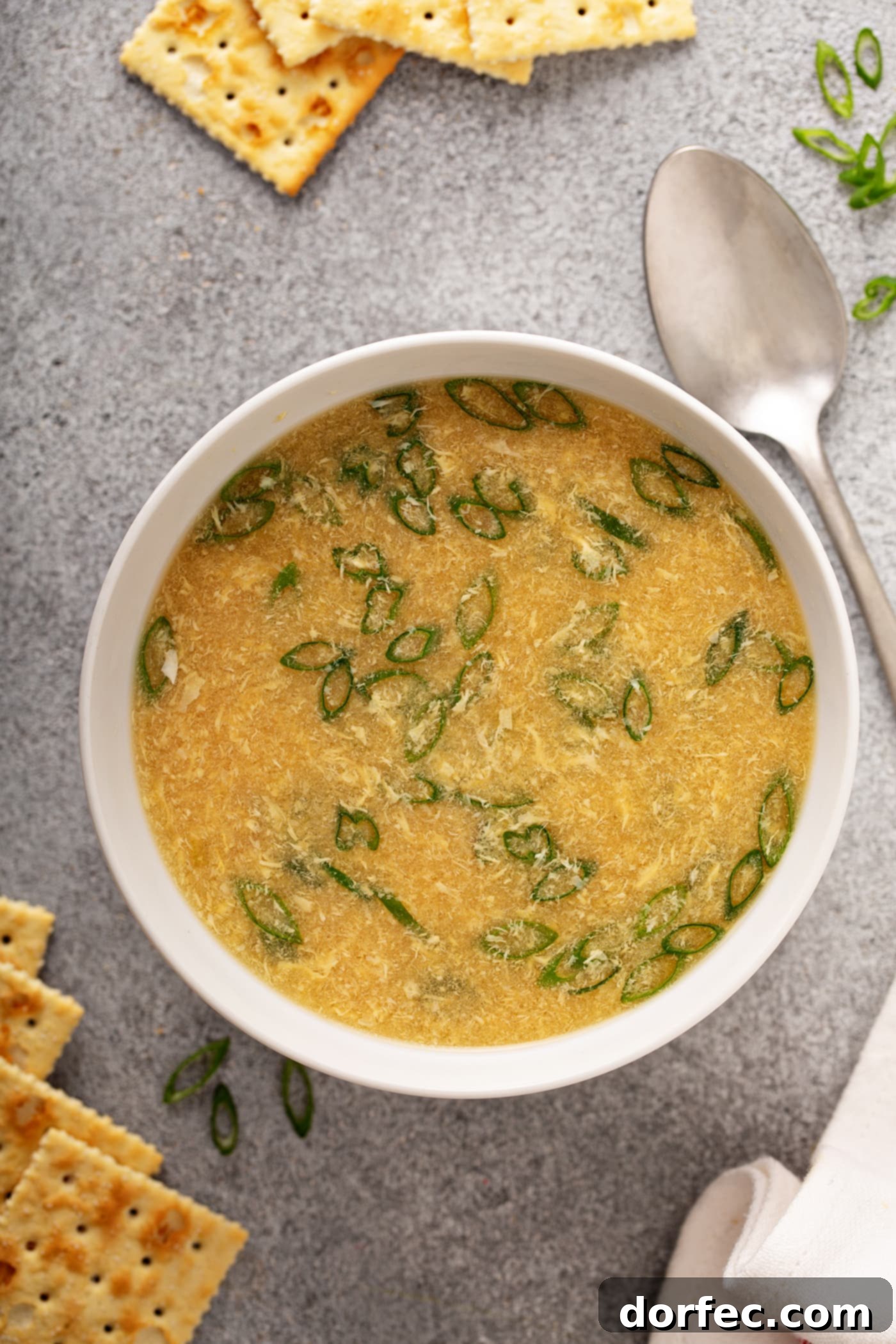 Easy Egg Drop Soup A bowl of egg drop soup with a spoon and three saltine crackers laying next to it. You can see bits of egg and sliced green peppers in the soup.