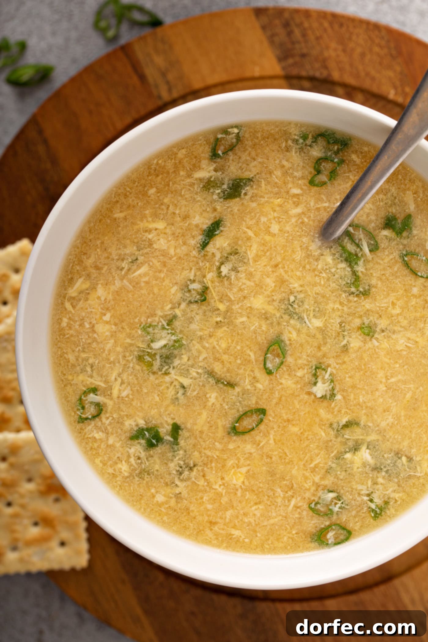 Easy Egg Drop Soup A close-up of a bowl of egg drop soup with sliced onions on top and saltine crackers in the background.