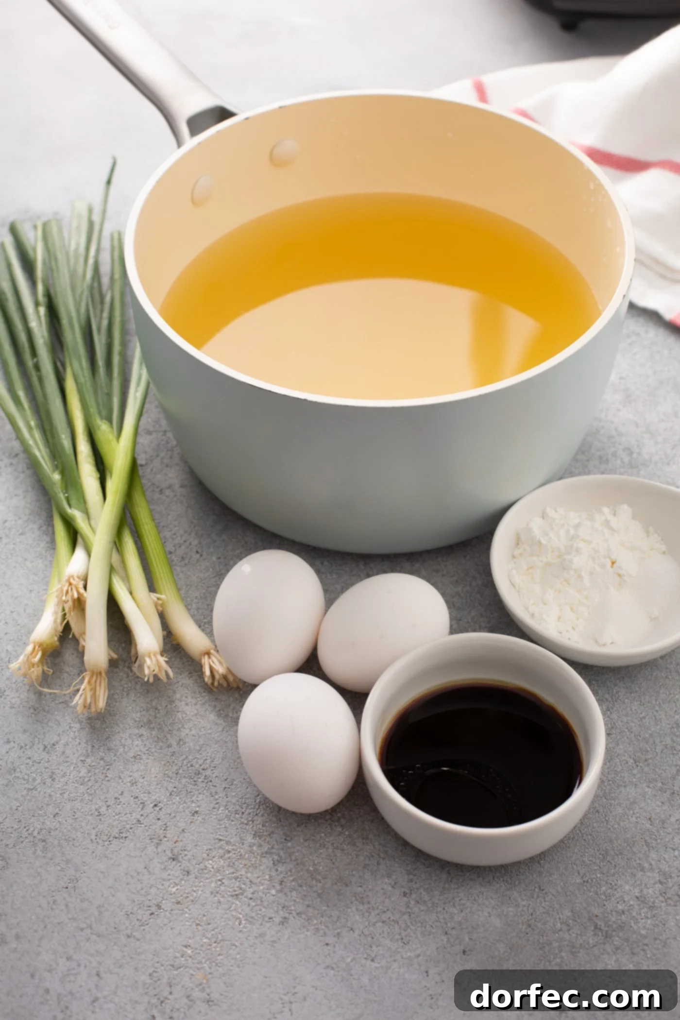 Easy Egg Drop Soup A table top with green onions, three whole eggs, soy sauce, a container of corn starch and a bowl of chicken broth.