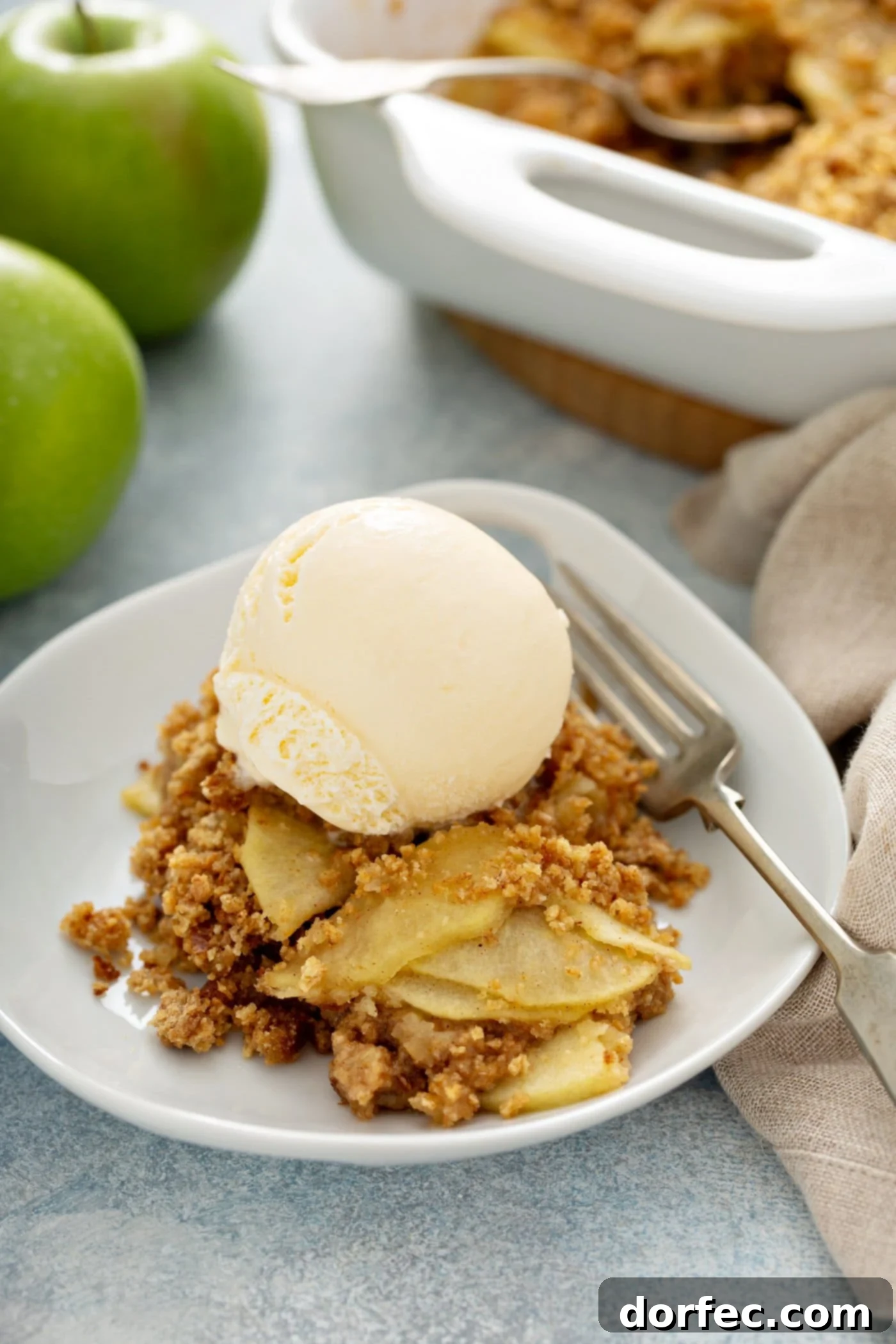 A serving of Apple Brown Betty in a glass dish, topped with a scoop of melting vanilla ice cream and a fresh sprig of mint. A fork rests on the white plate next to the dessert. In the soft-focus background, green apples, a baking dish with more Apple Brown Betty, and a linen napkin complete the cozy scene.