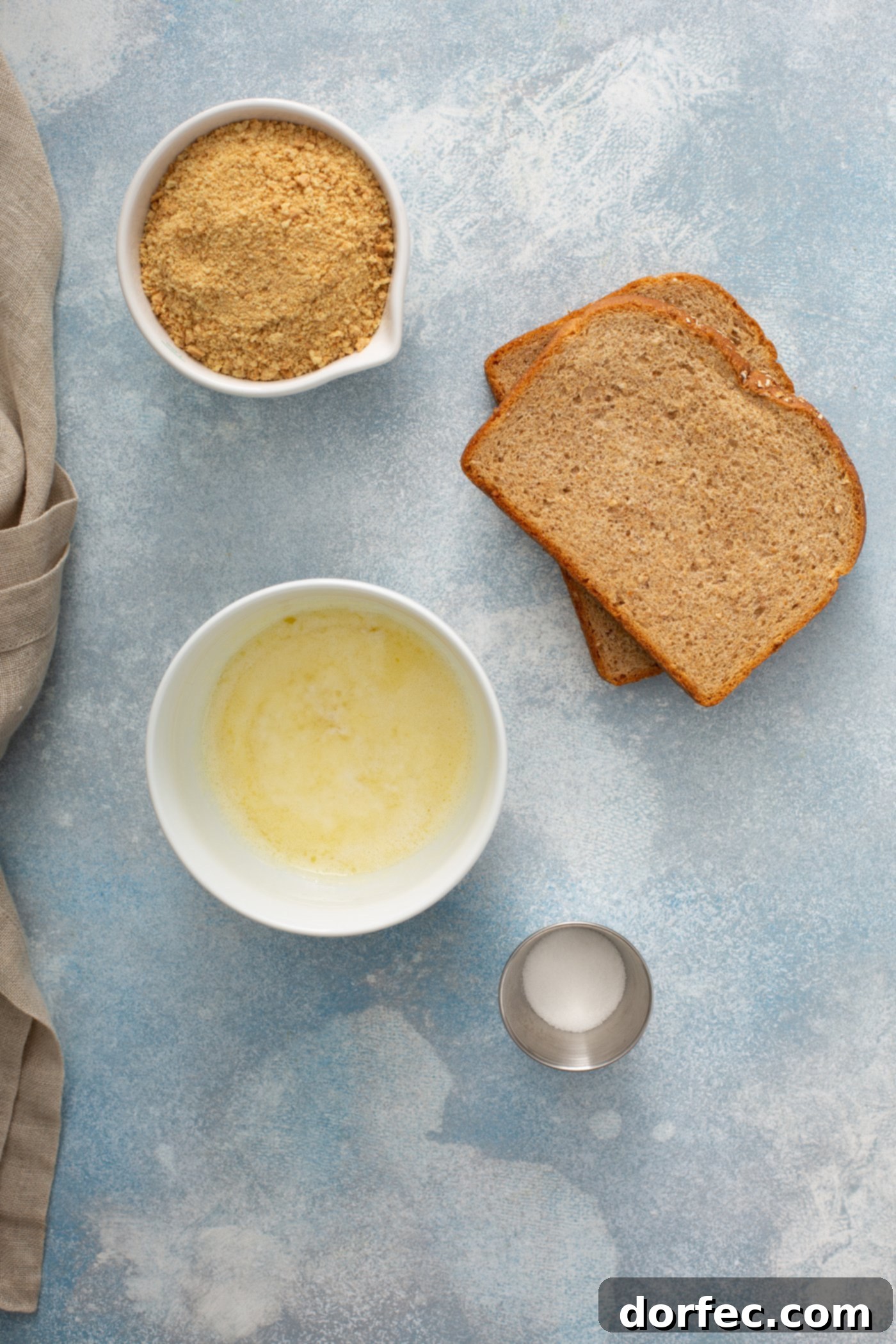 Ingredients for the Apple Brown Betty topping displayed on a rustic wooden tabletop: two slices of brown bread, a small bowl of melted butter, and separate containers of sugar and graham cracker crumbs.
