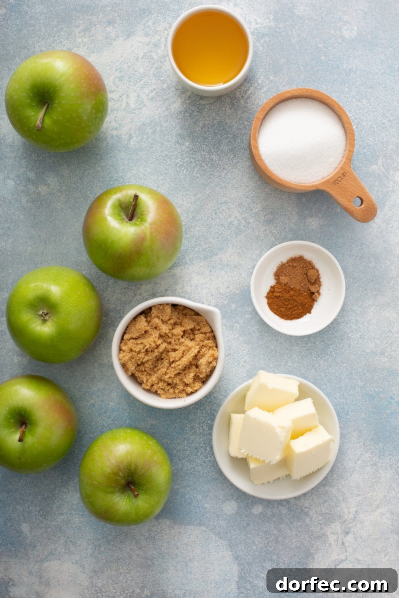 A colorful arrangement of ingredients for the Apple Brown Betty filling: five vibrant green apples, alongside small bowls containing cubed butter, ground cinnamon, bread crumbs, and granulated sugar.