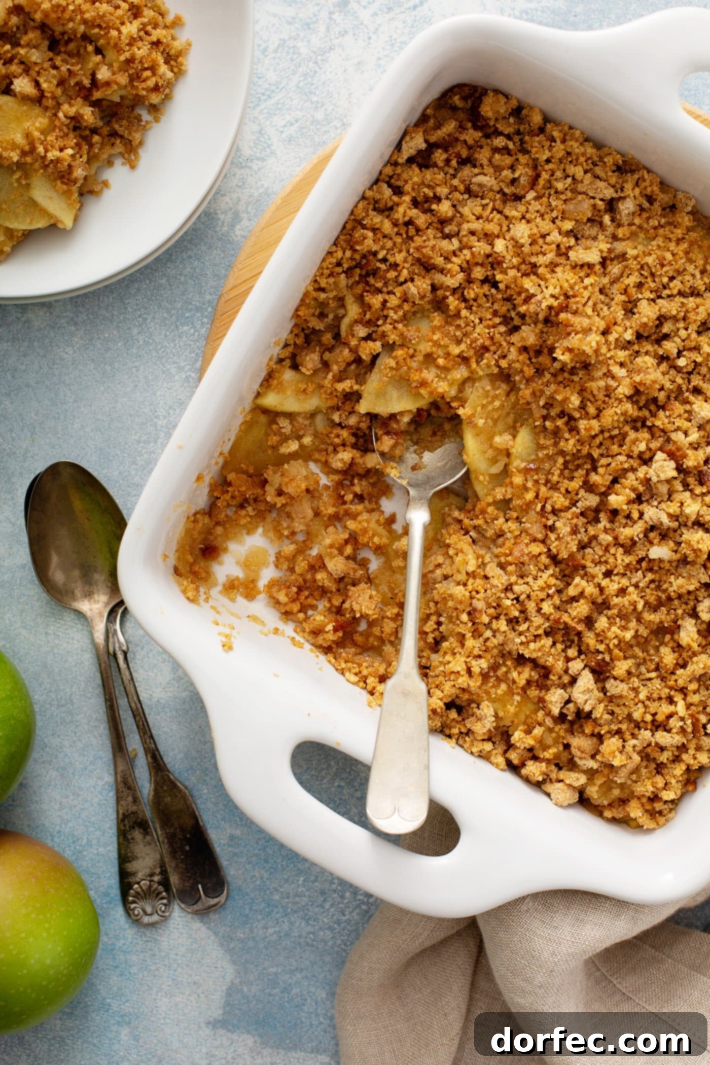A white ceramic baking dish brimming with a golden-brown Apple Brown Betty, featuring visible sliced apples beneath a crispy breadcrumb topping. A serving spoon is nestled in the dessert, with additional servings in dessert dishes and various spoons blurred in the background.