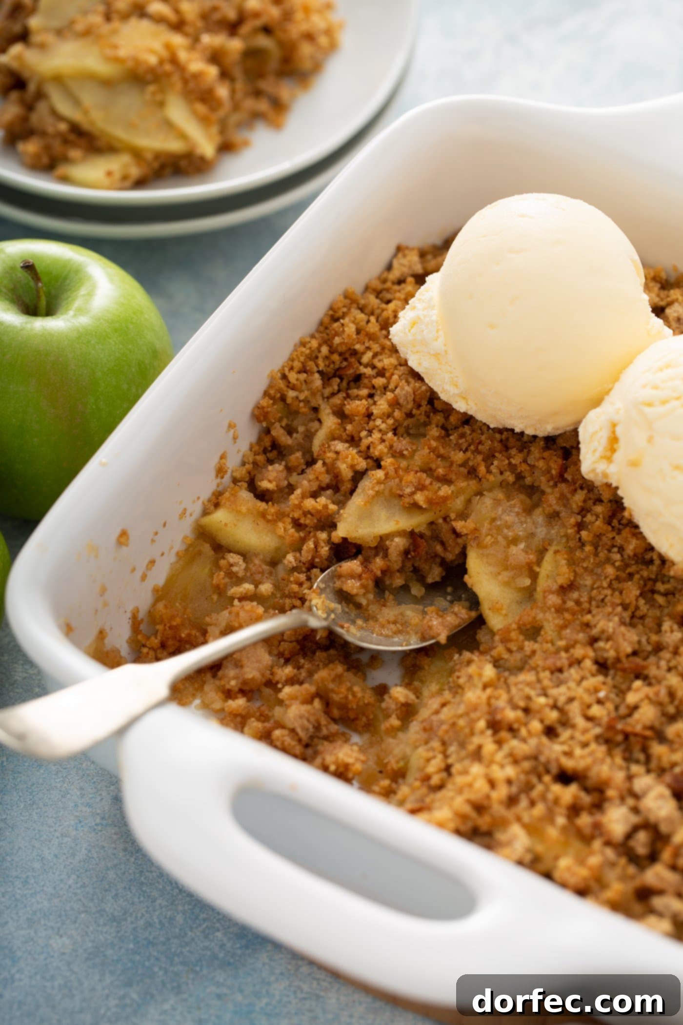 A white baking dish filled with a golden-brown Apple Brown Betty, crowned with two scoops of creamy vanilla ice cream. A single green apple rests beside the dish, with other dessert plates subtly blurred in the background.