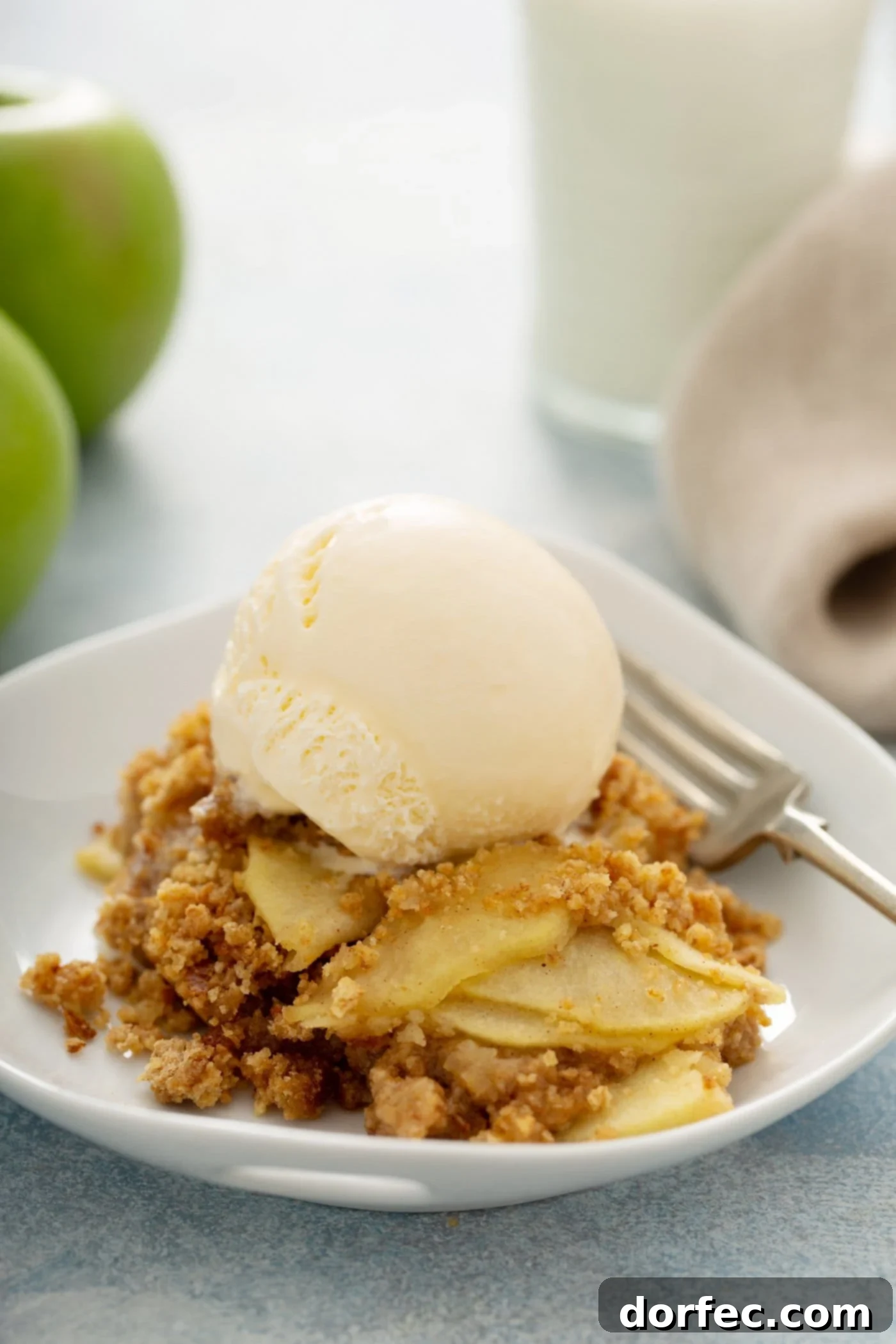 A close-up shot of Apple Brown Betty on a dessert plate, showcasing tender apple slices beneath a rich, brown breadcrumb topping. A single scoop of vanilla ice cream adorns the dessert, with a fork positioned neatly beside it on the plate. Some fresh green apples are visible in the background, hinting at the key ingredient.