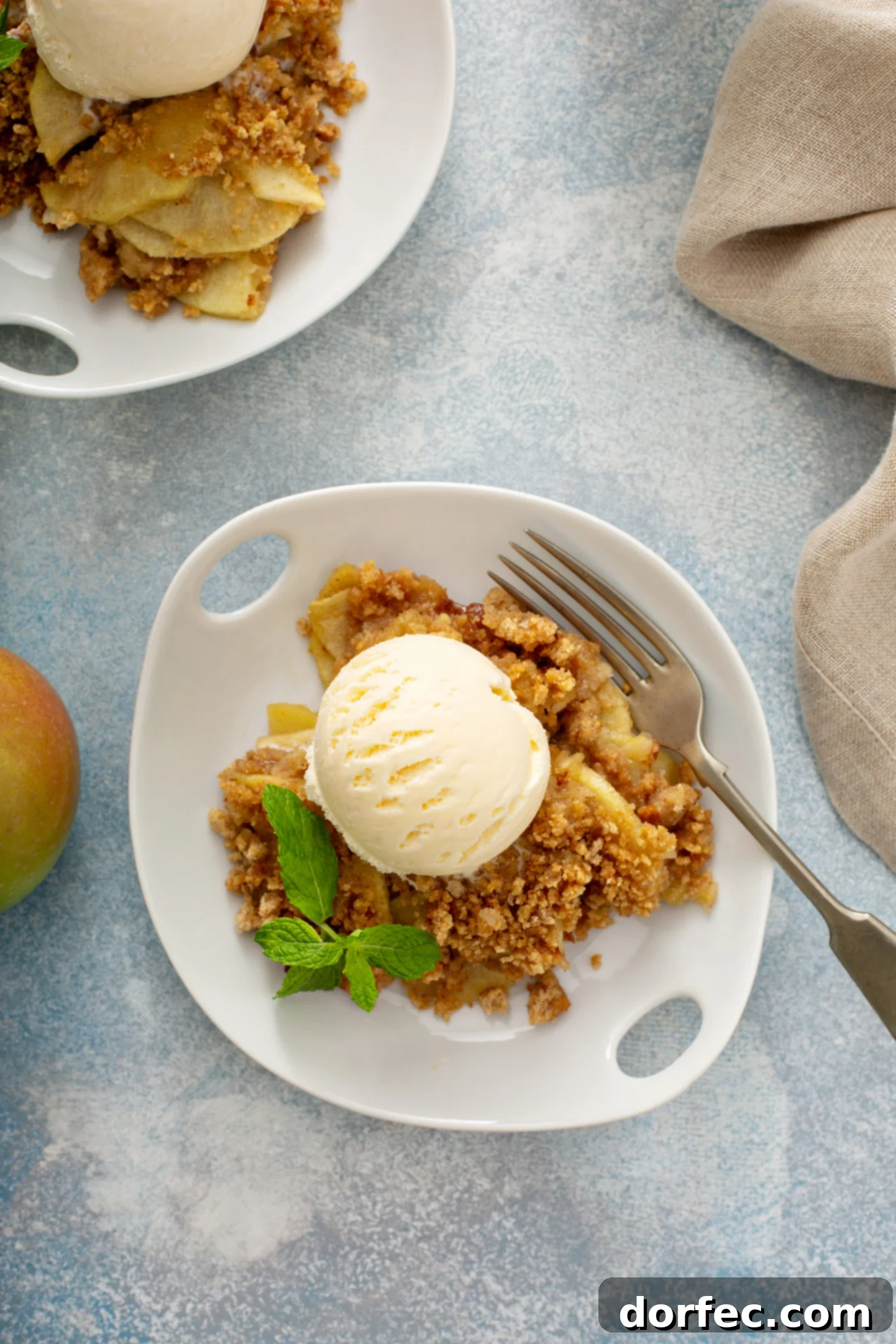A close-up of a single serving of Apple Brown Betty on a pristine white dessert plate. The warm apple dessert is elegantly topped with a generous scoop of vanilla ice cream, and a delicate sprig of fresh peppermint adds a touch of color. A second serving is softly blurred in the background, creating a cozy and inviting scene.