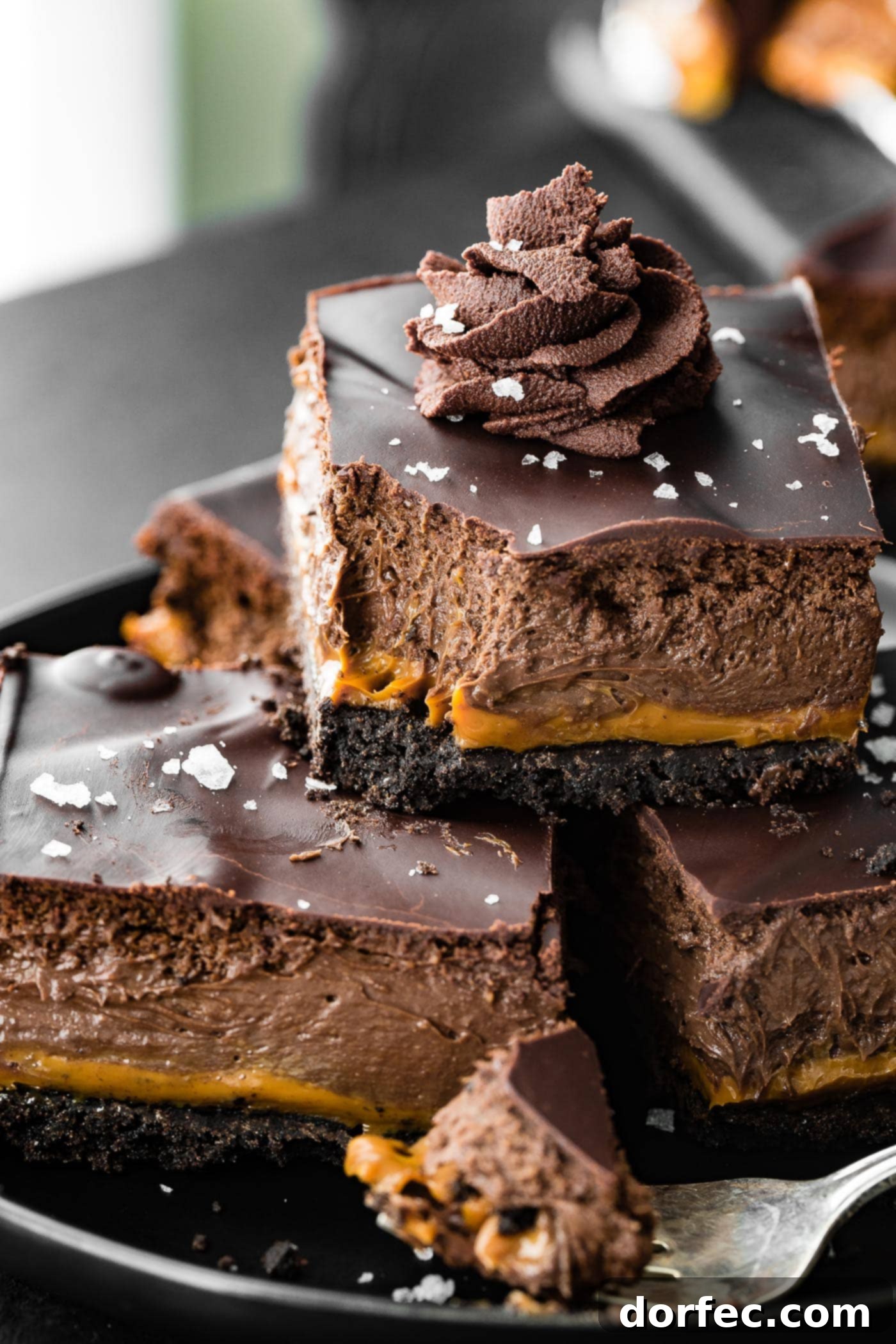A bite is taken out of a layered Dulce de Leche Dessert Cream Cheese Bar with whipped ganache on top. The dessert is layered with oreo crust, creamy caramel, chocolate cheesecake filling, and a chocolate ganache topping, garnished with sea salt.