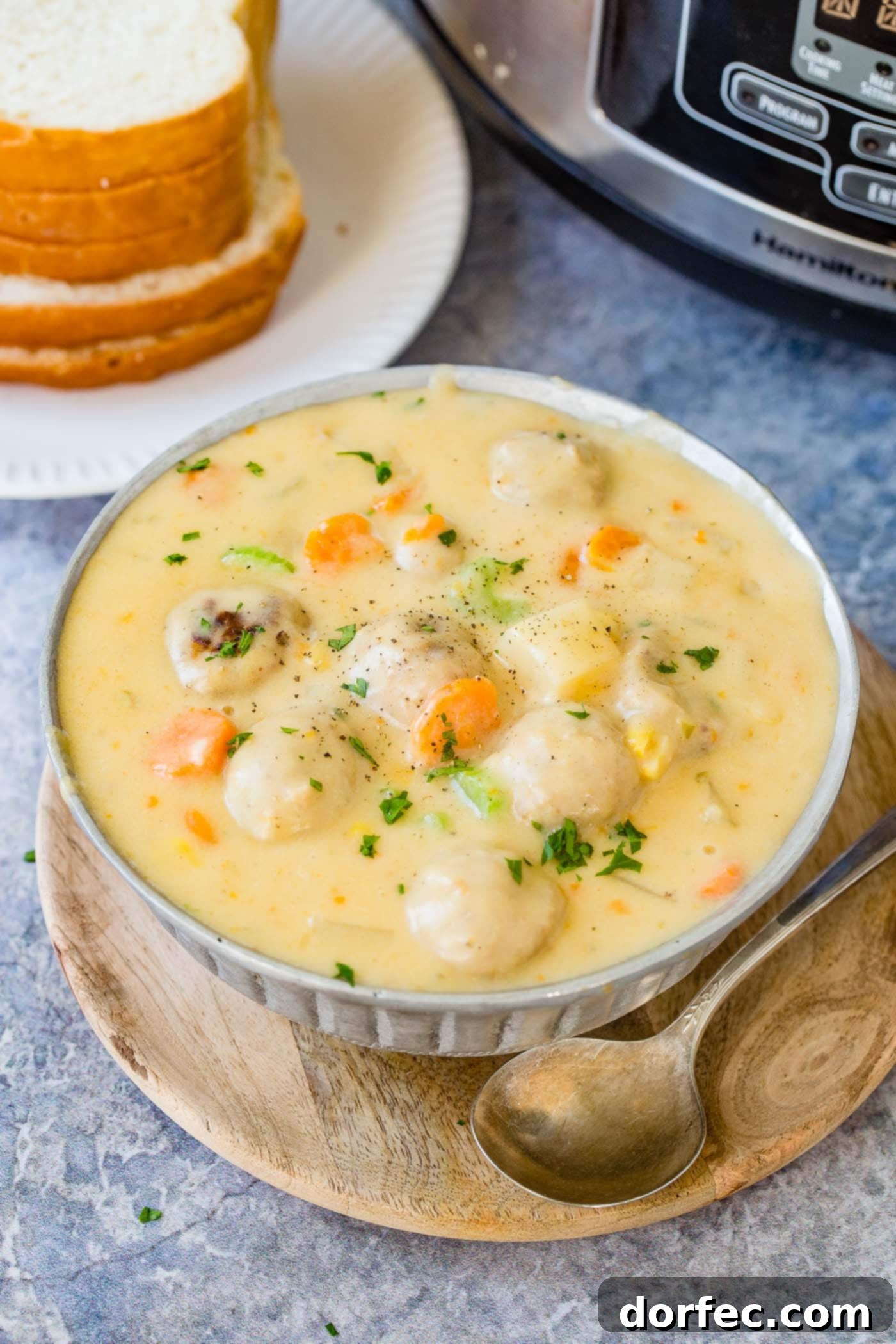 A glistening metal bowl filled with warm, cheesy meatball soup, garnished with green herbs, with slices of soft white bread in the background, ready for a comforting dip.