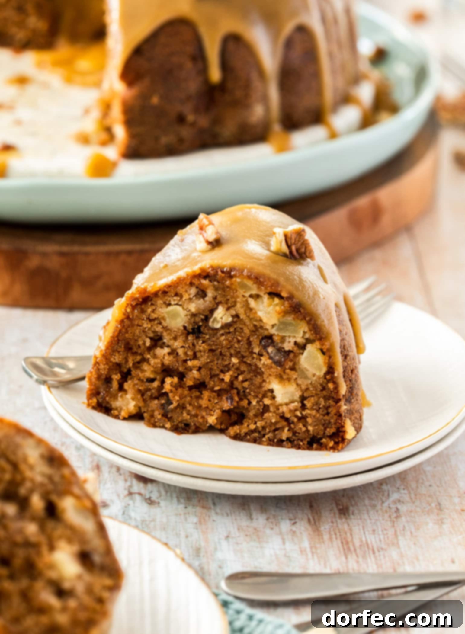 A perfect slice of Pecan Apple Bundt Cake with caramel icing, served on a white plate, with the remainder of the cake in the background, showcasing its moist texture.