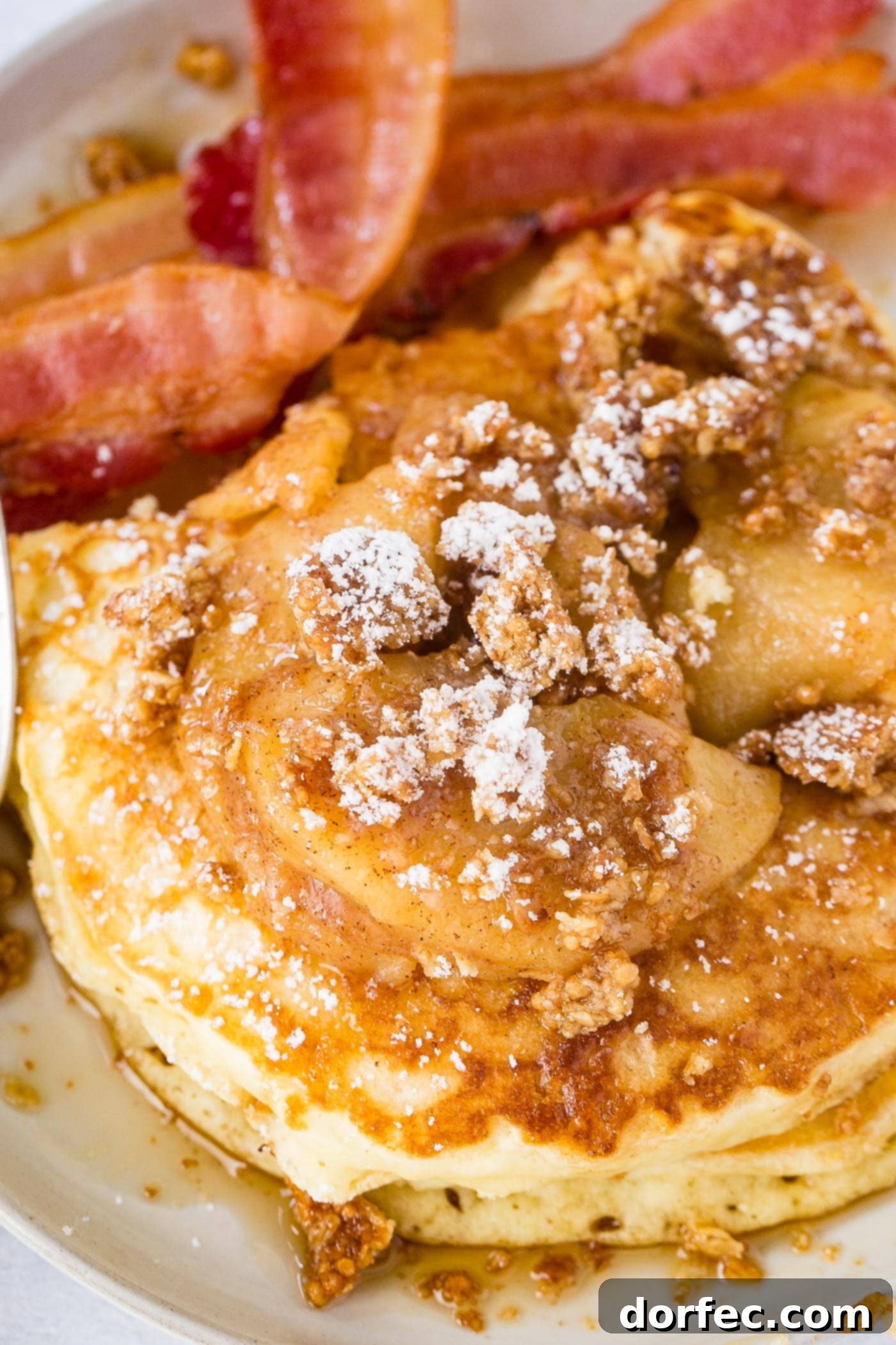 A plate stacked high with fluffy buttermilk pancakes, adorned with glistening roasted cinnamon apples, crumbled granola, a dusting of powdered sugar, and a generous pour of maple syrup. A side of crispy bacon completes this delightful breakfast scene.