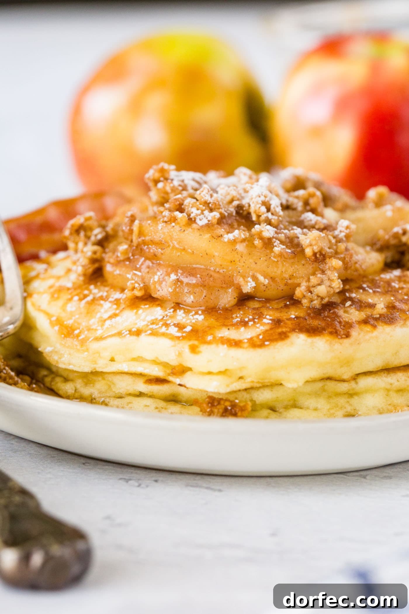 A close-up shot of a stack of golden, fluffy pancakes generously topped with roasted cinnamon apples and crunchy granola. A light dusting of powdered sugar and a rich drizzle of maple syrup complete the presentation. Crispy bacon is visible on the side.
